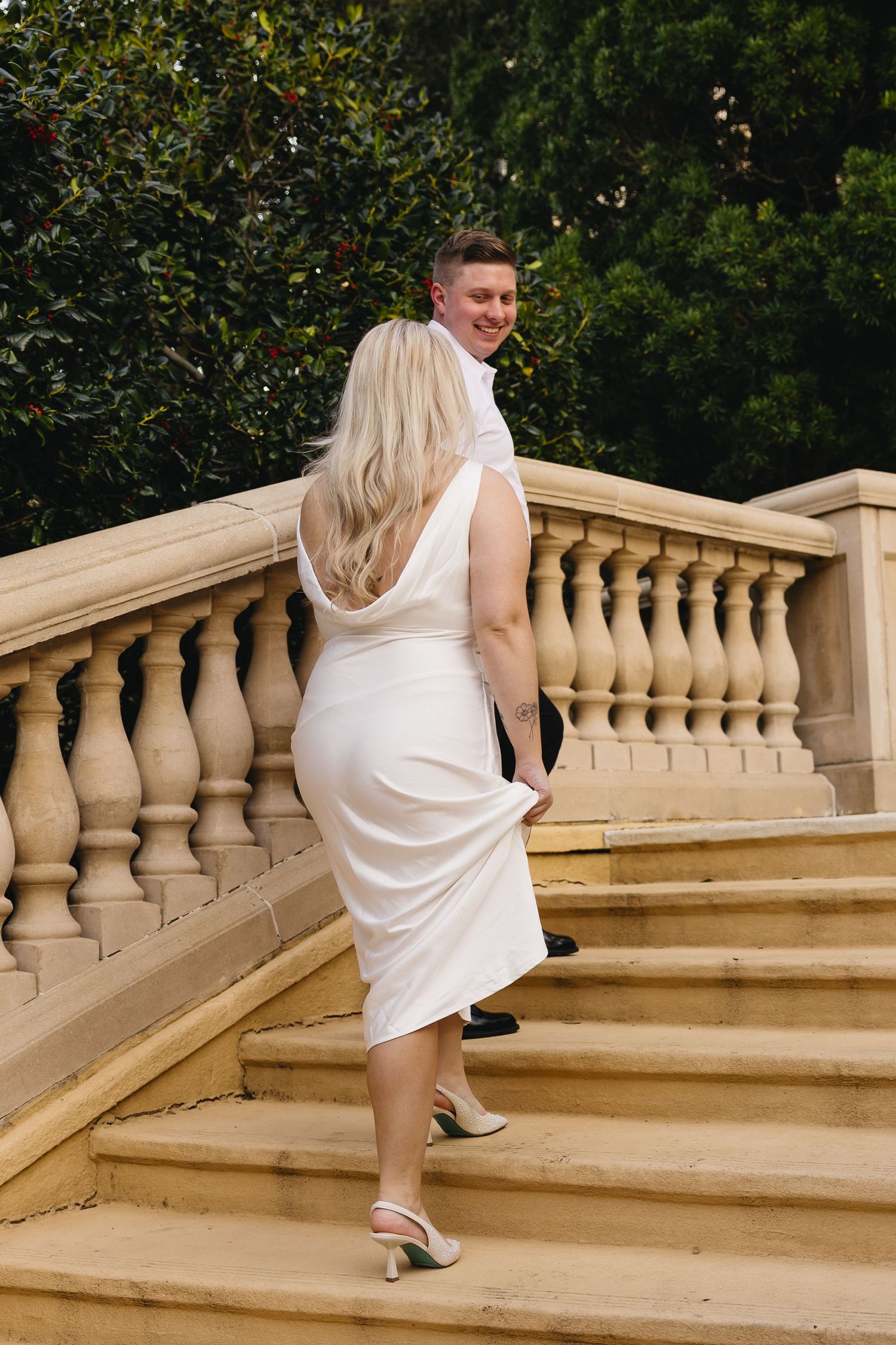 Bride in white satin dress leads groom up elegant stone staircase with balustrade surrounded by lush greenery.