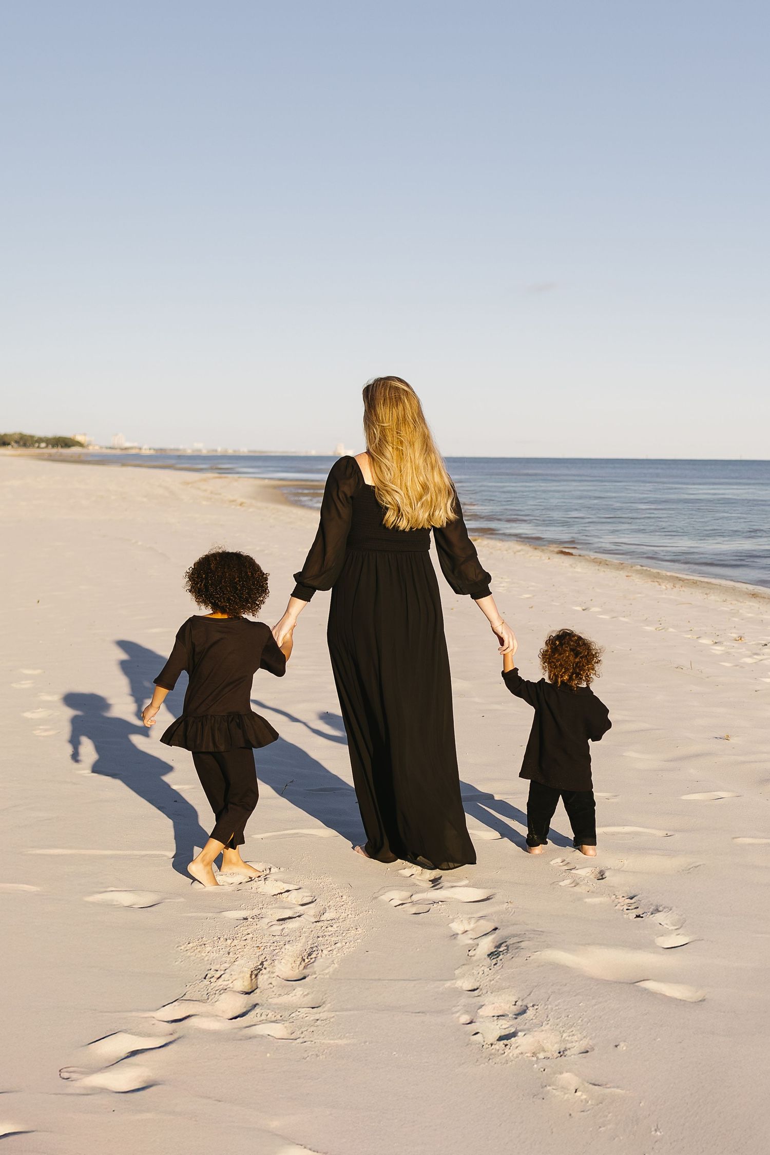 A mother in a black dress walks hand-in-hand with two young children on a sandy beach.