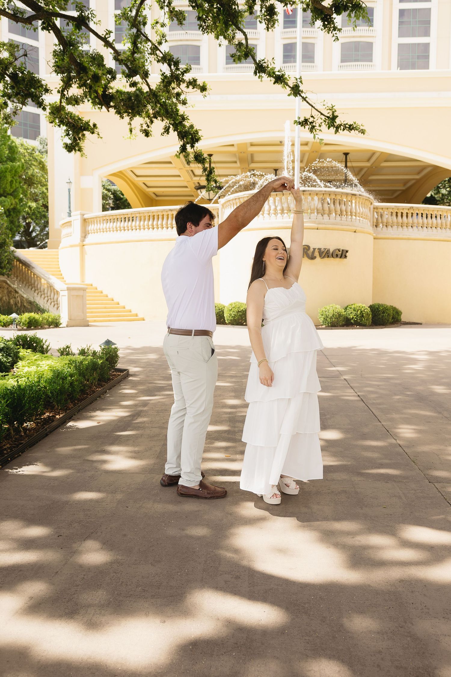 Man twirling pregnant woman in a white dress outside the Beau Rivage resort on a sunny day.