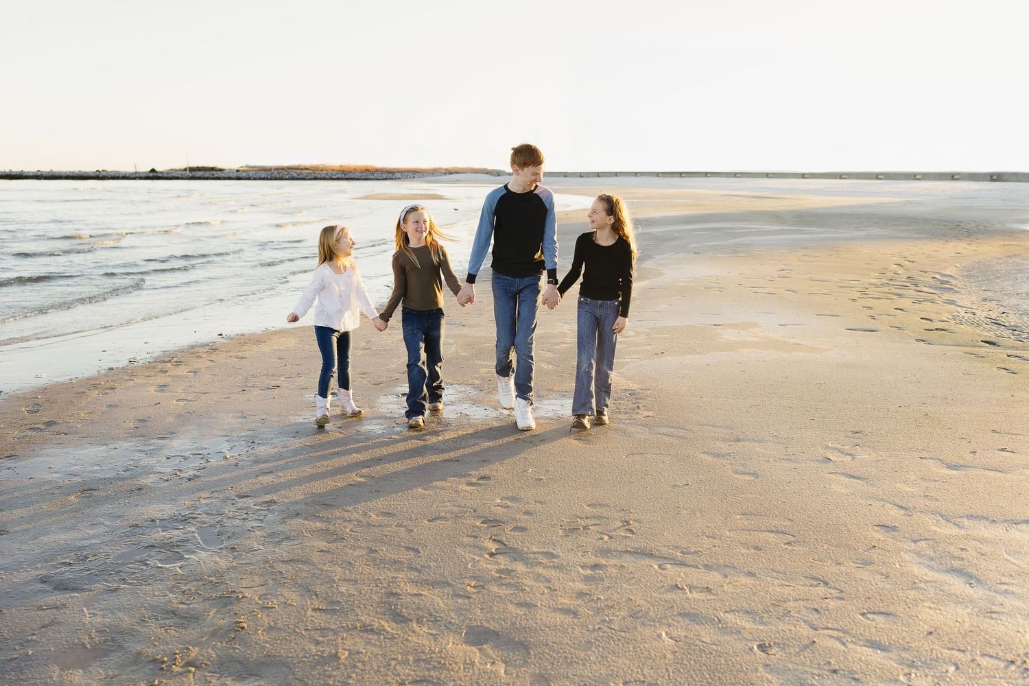 Four siblings hold hands and walk together along a sunlit shoreline on a sandy beach.