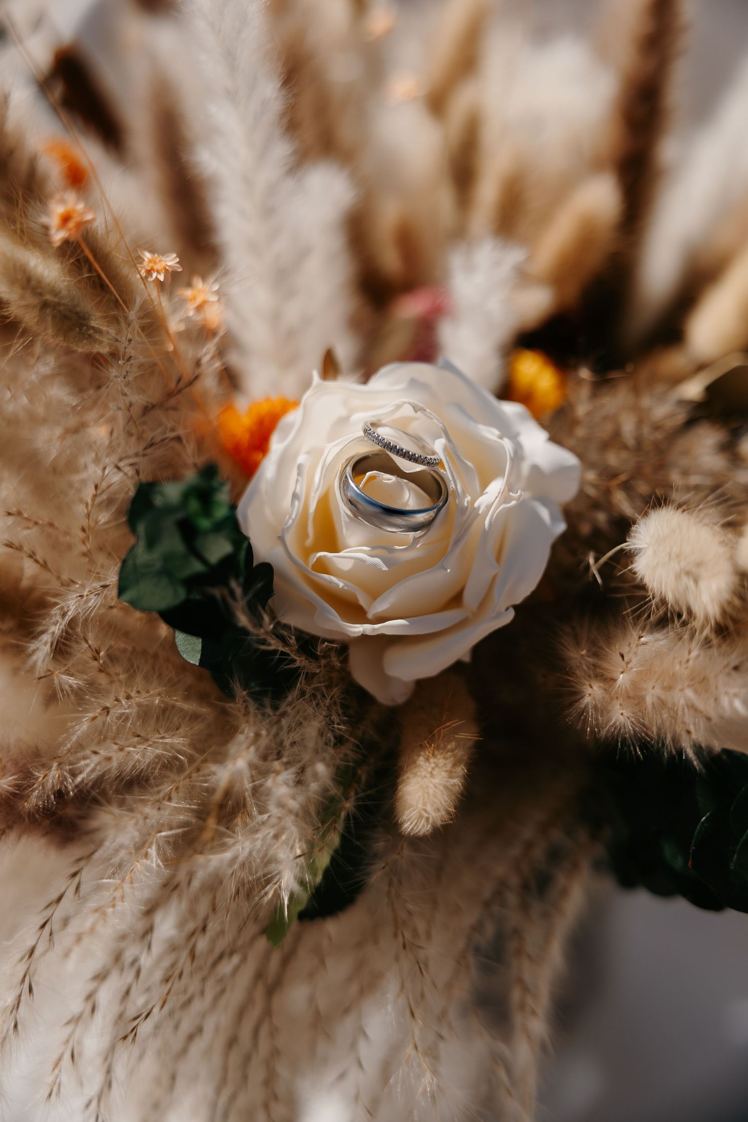 Two silver wedding rings resting on a white rose in a boho dried floral bouquet with pampas grass.