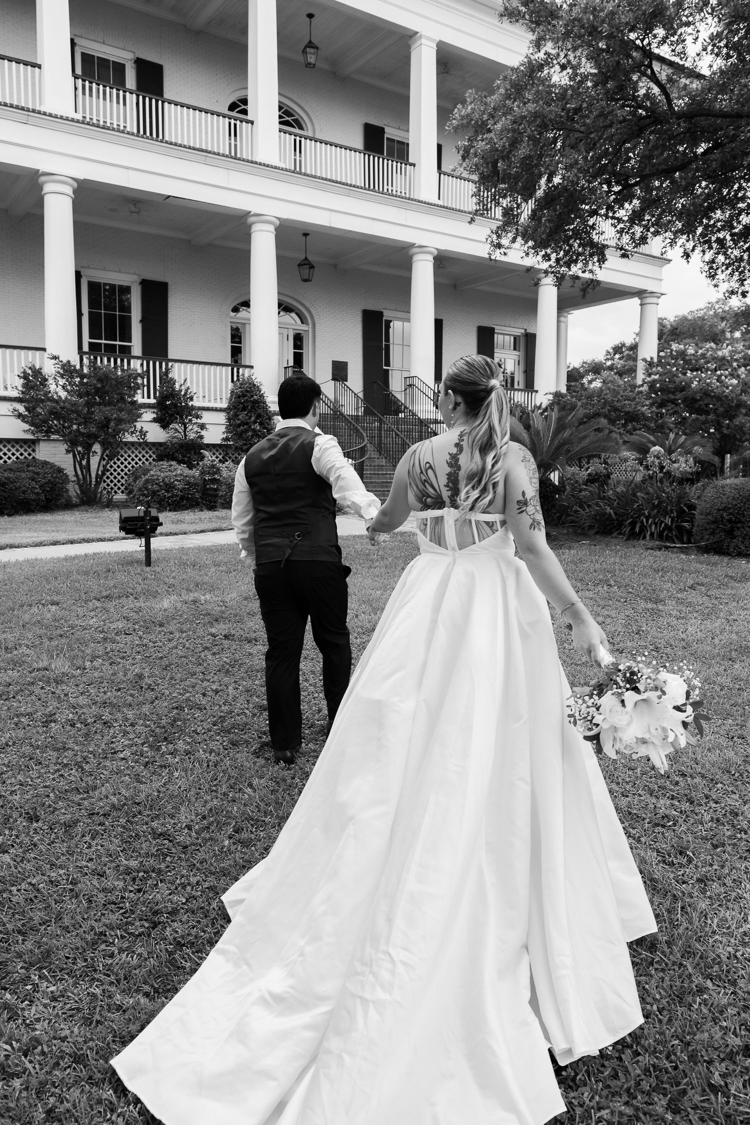 Black and white photo of bride and groom walking toward a grand Southern plantation home.