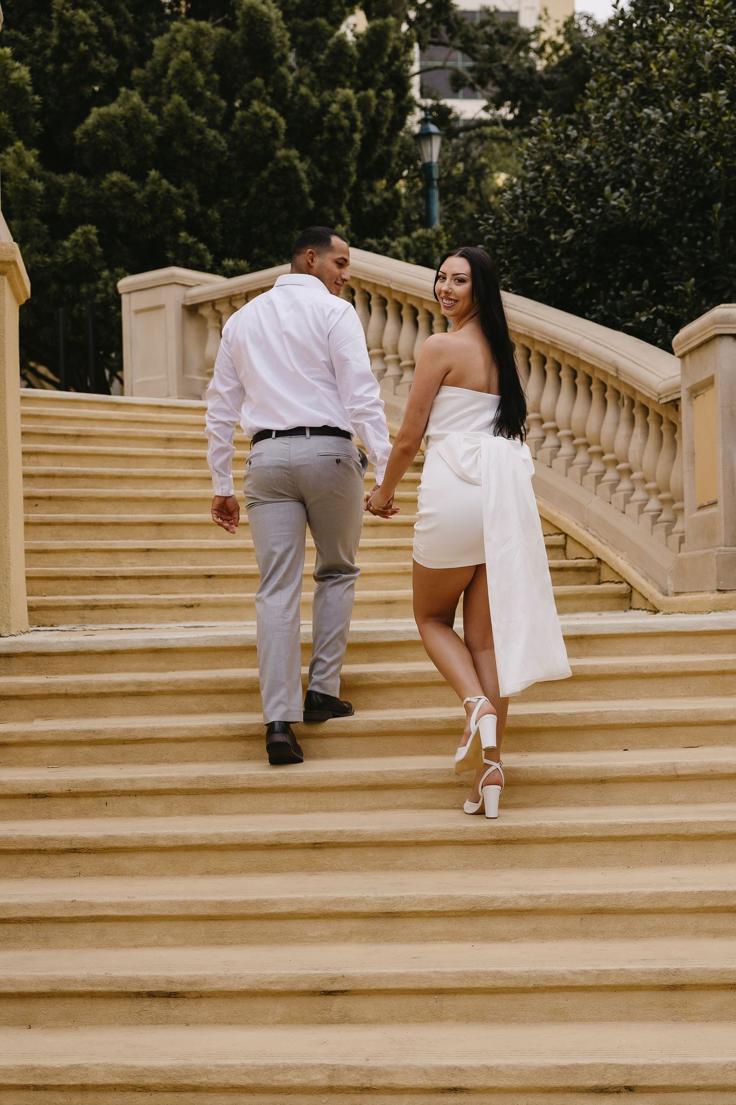 Couple holding hands walking up grand stone staircase, woman in white strapless mini dress glances back smiling.
