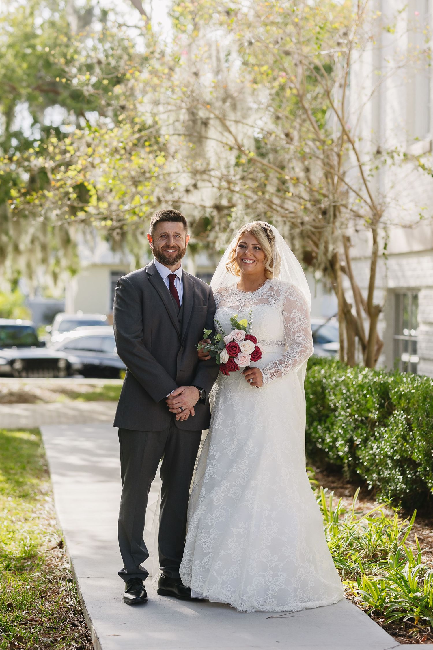 Smiling bride and groom pose on a sunny sidewalk, bride holding red and white bouquet in lace gown with veil.