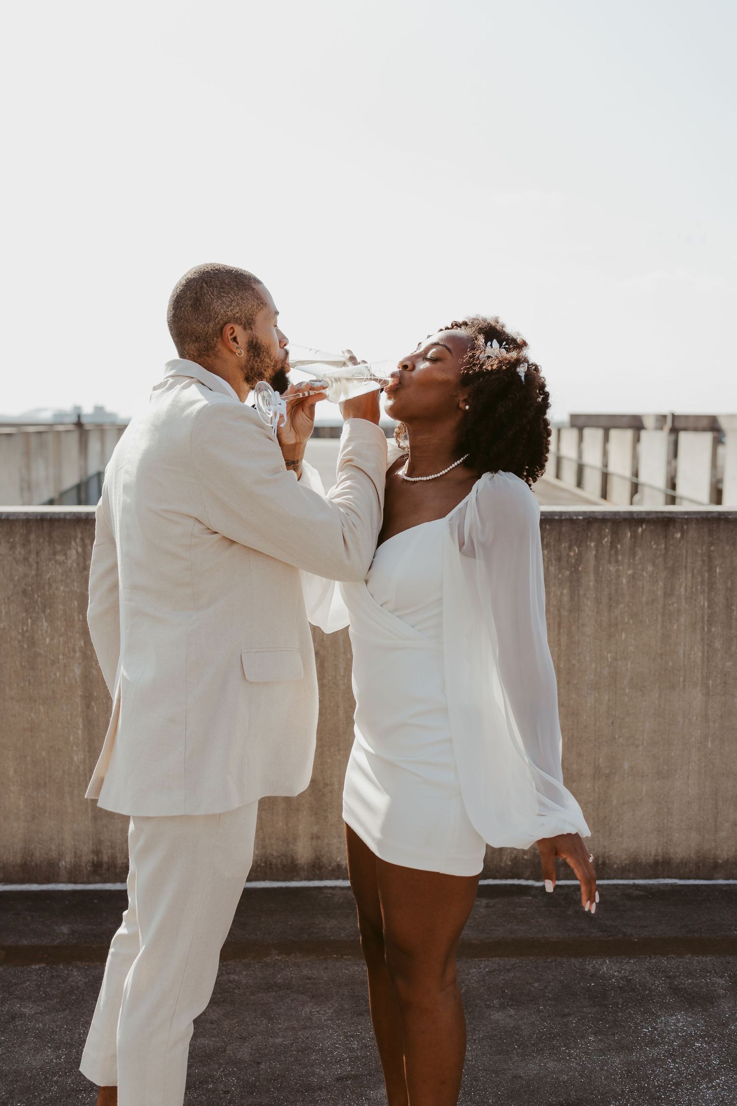 Bride and groom in matching white outfits sharing a champagne drink together on a sunny rooftop.