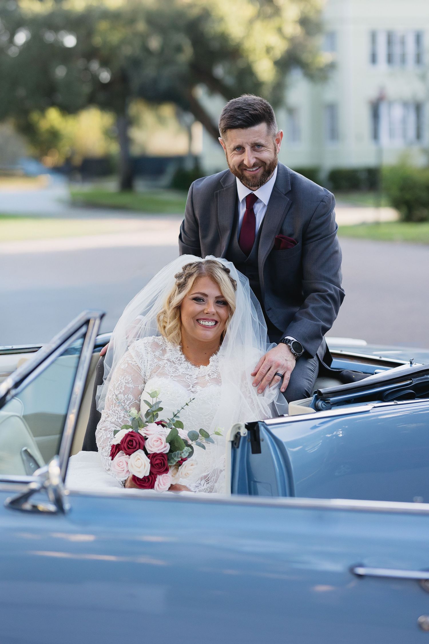 Smiling bride in lace gown holding red and white bouquet with groom beside vintage convertible.