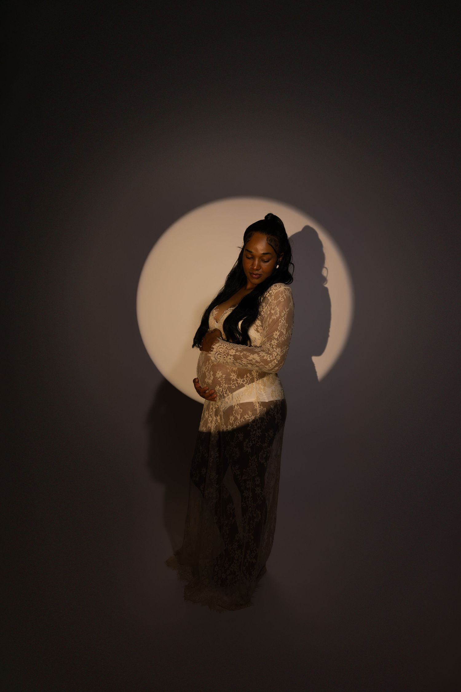 Pregnant woman in lace gown standing in dramatic circular spotlight against dark background.