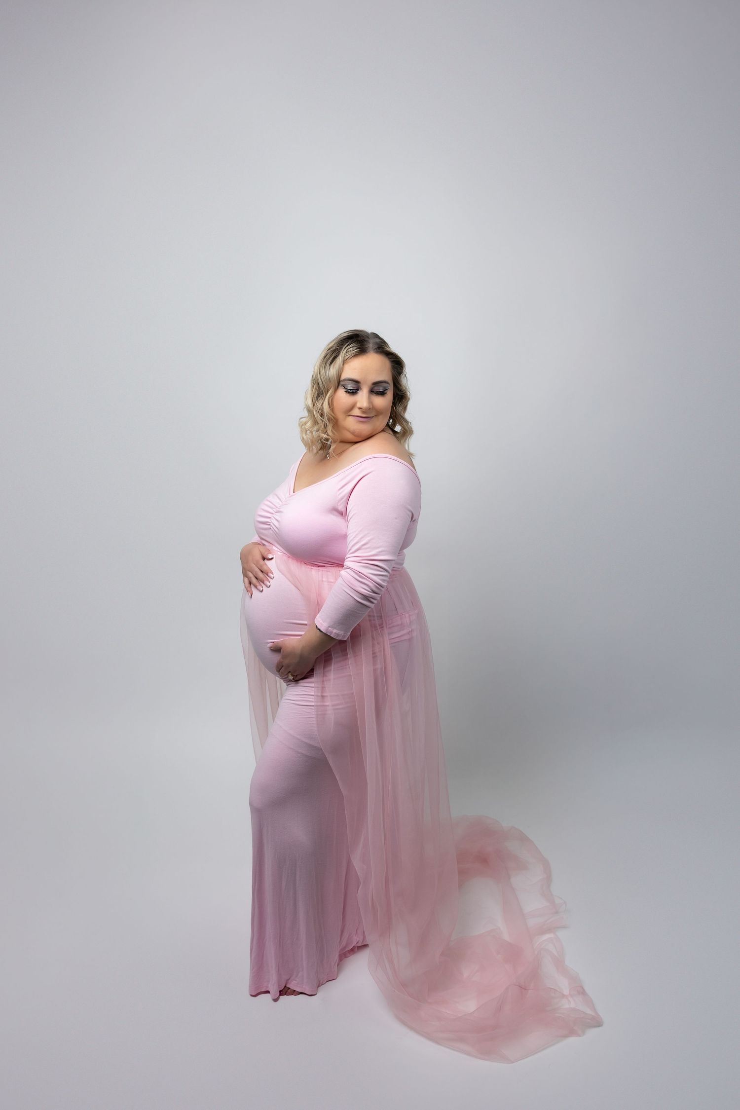 Pregnant woman in pink bodycon dress with tulle train poses gracefully in studio.