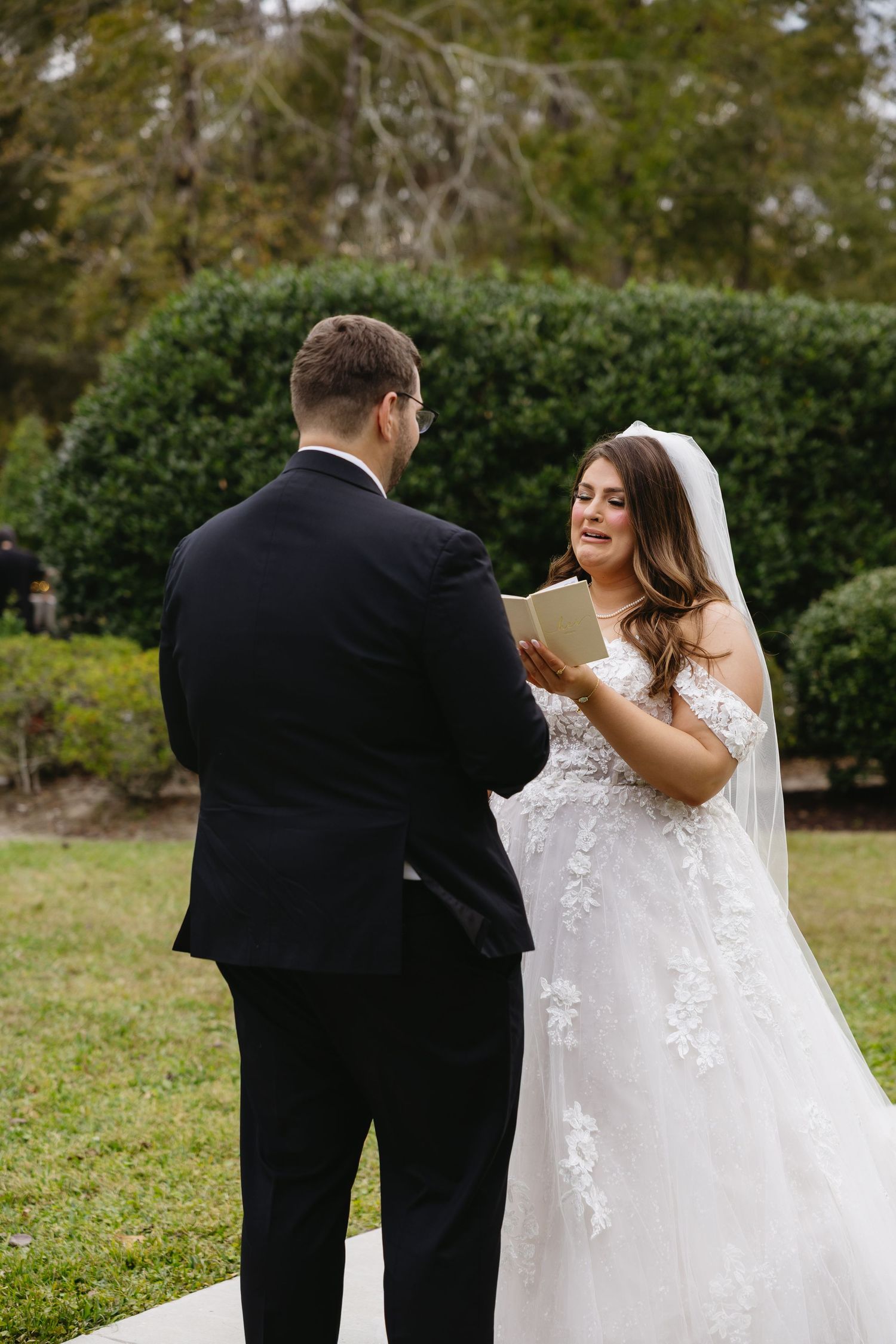 Bride reads vows from a card to groom during outdoor wedding ceremony surrounded by greenery.