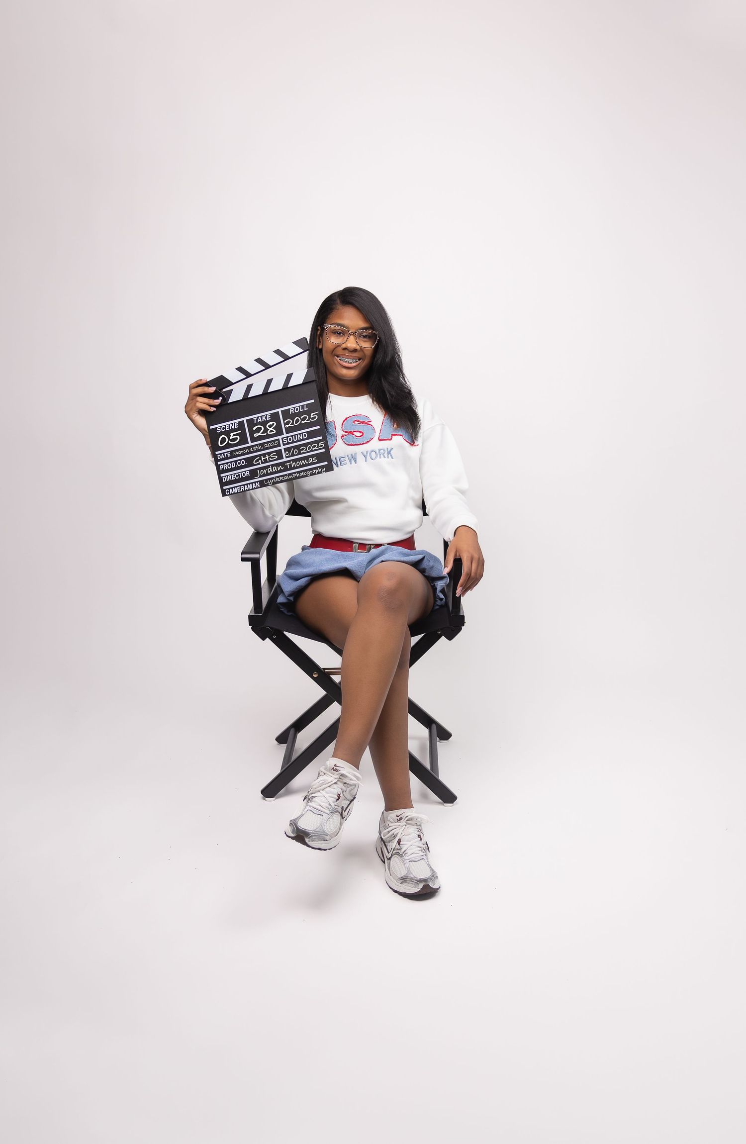 Smiling young woman holding film clapperboard seated in directors chair in studio portrait.