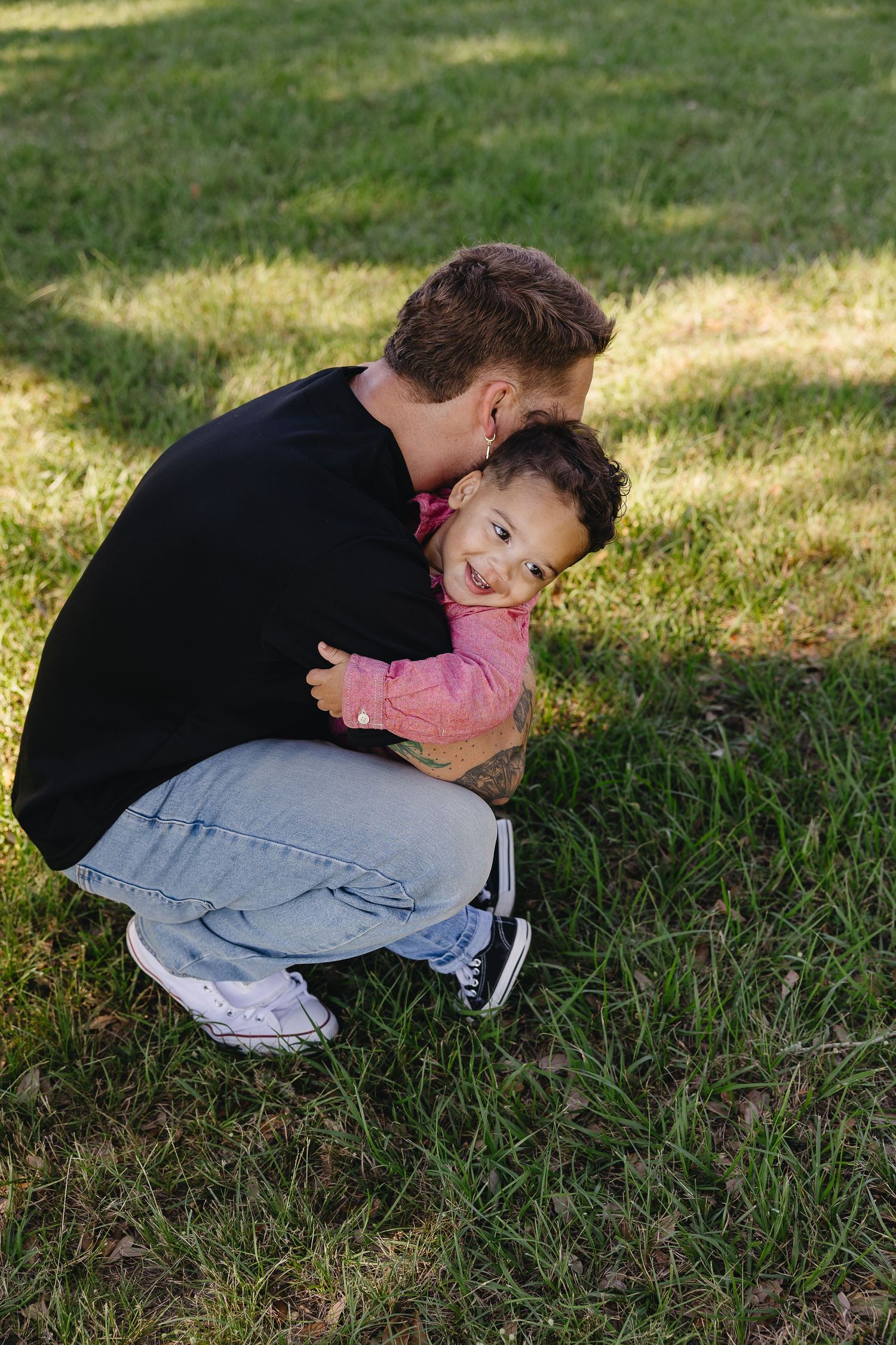 Tattooed father crouching on grass hugging smiling toddler in pink shirt in sunny park setting.