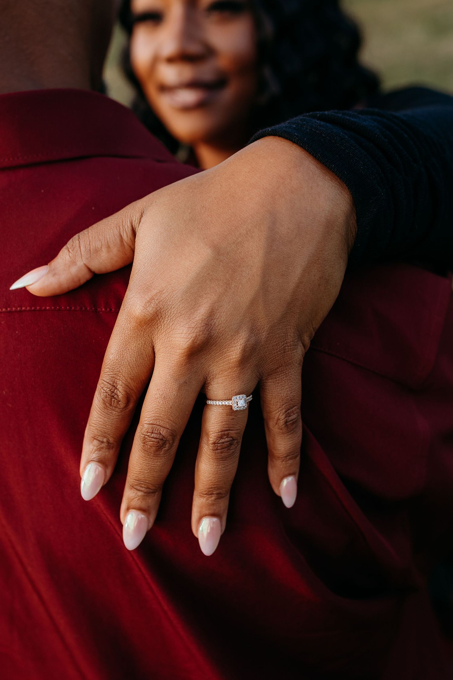 Close-up of a smiling Black woman showing off her diamond engagement ring on her partner's arm.