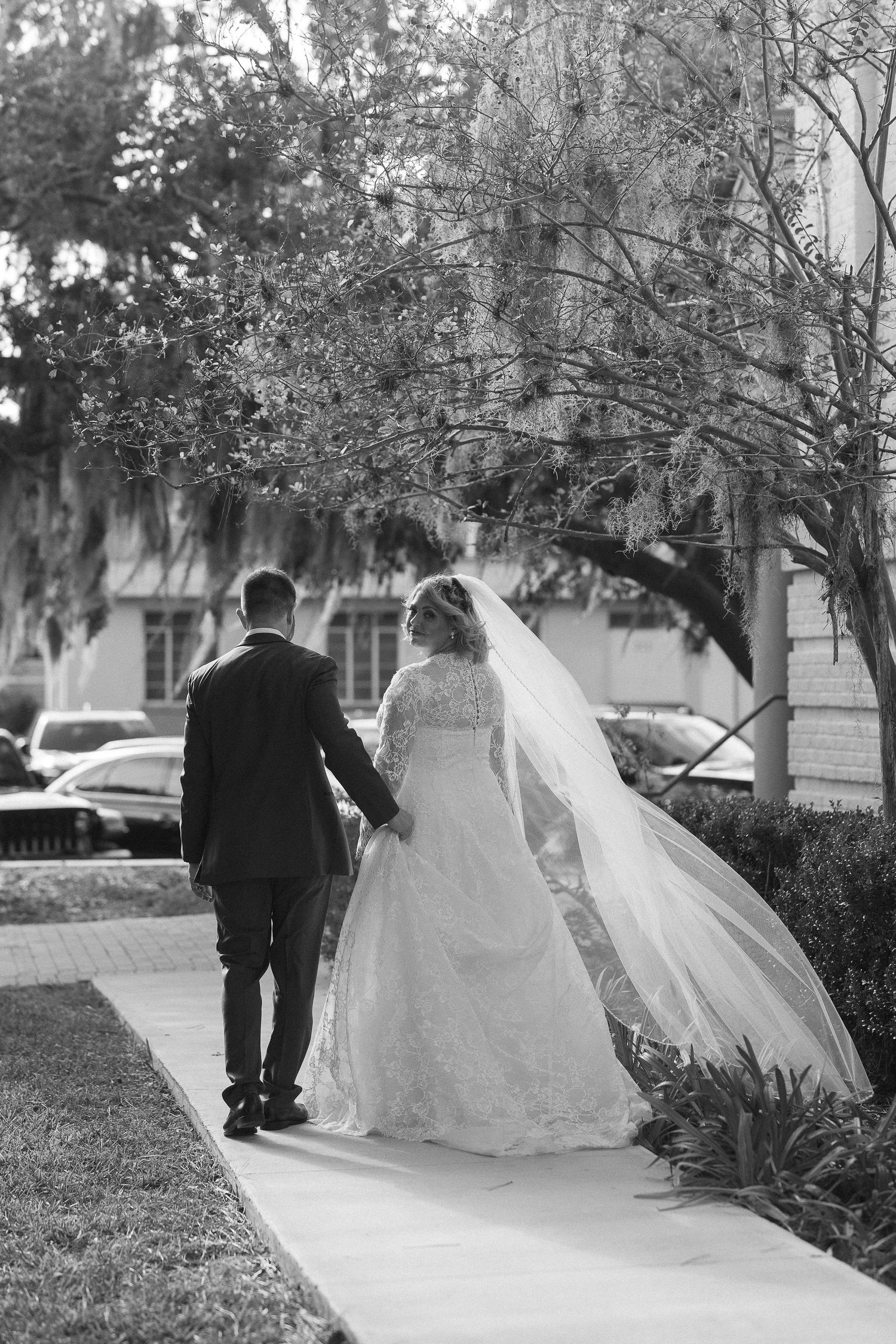 Bride and groom walking hand in hand on sidewalk in black and white wedding photo.