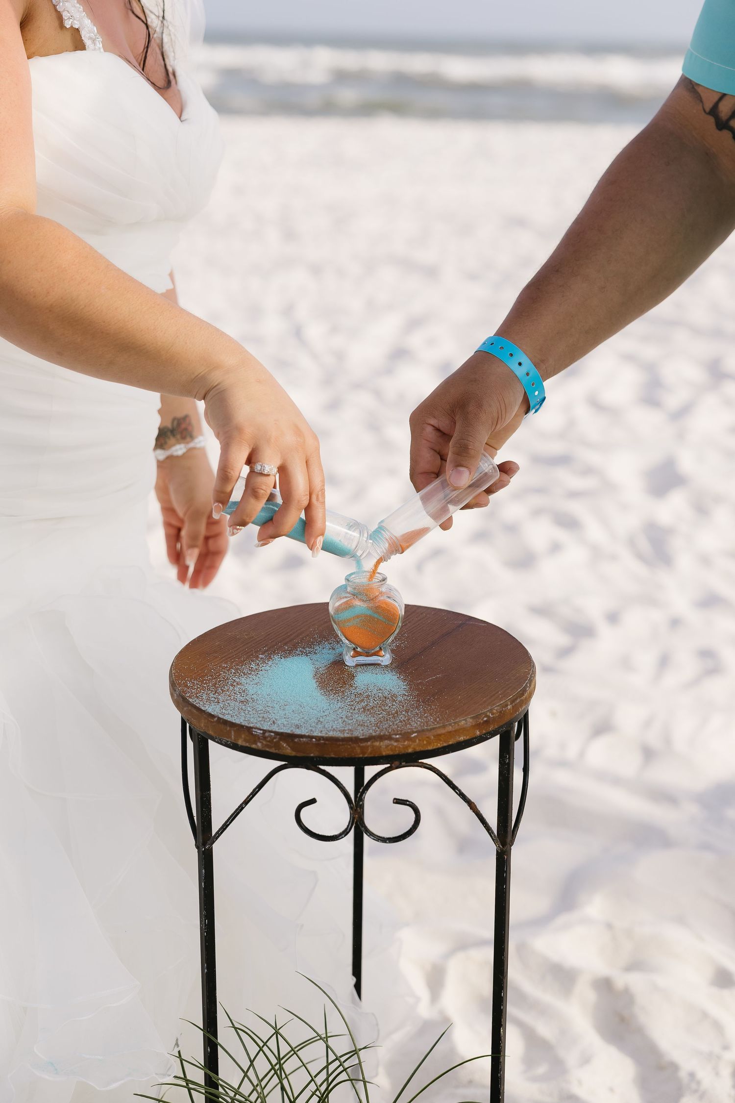 Bride and groom pouring colored sand into glass vase during beach wedding unity ceremony.