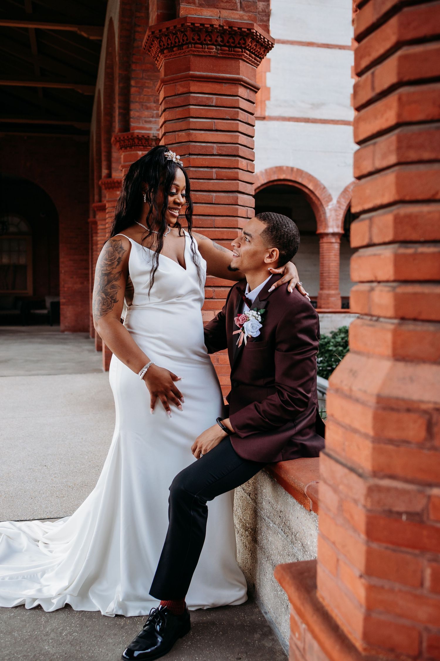 Groom in burgundy suit seated on brick ledge gazing at bride in white gown.