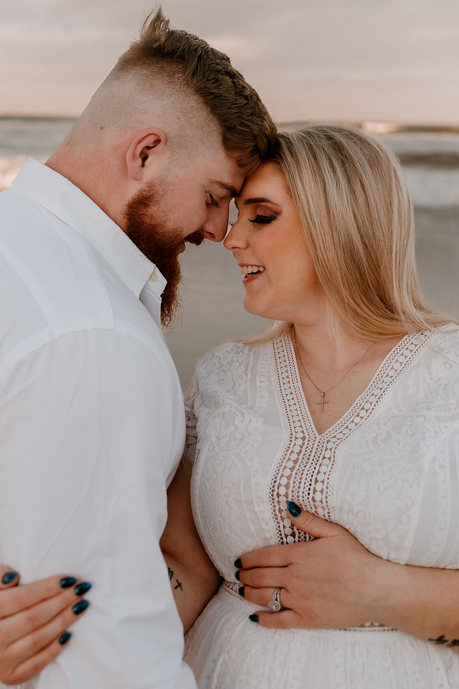 Close-up of smiling pregnant couple touching foreheads on beach, woman in white lace dress, golden hour light.