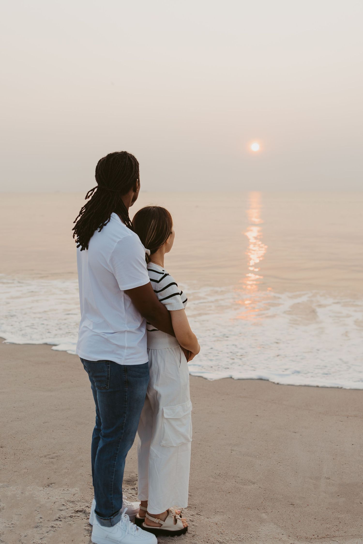 Interracial couple embracing while watching a golden sunset over the ocean on the beach.