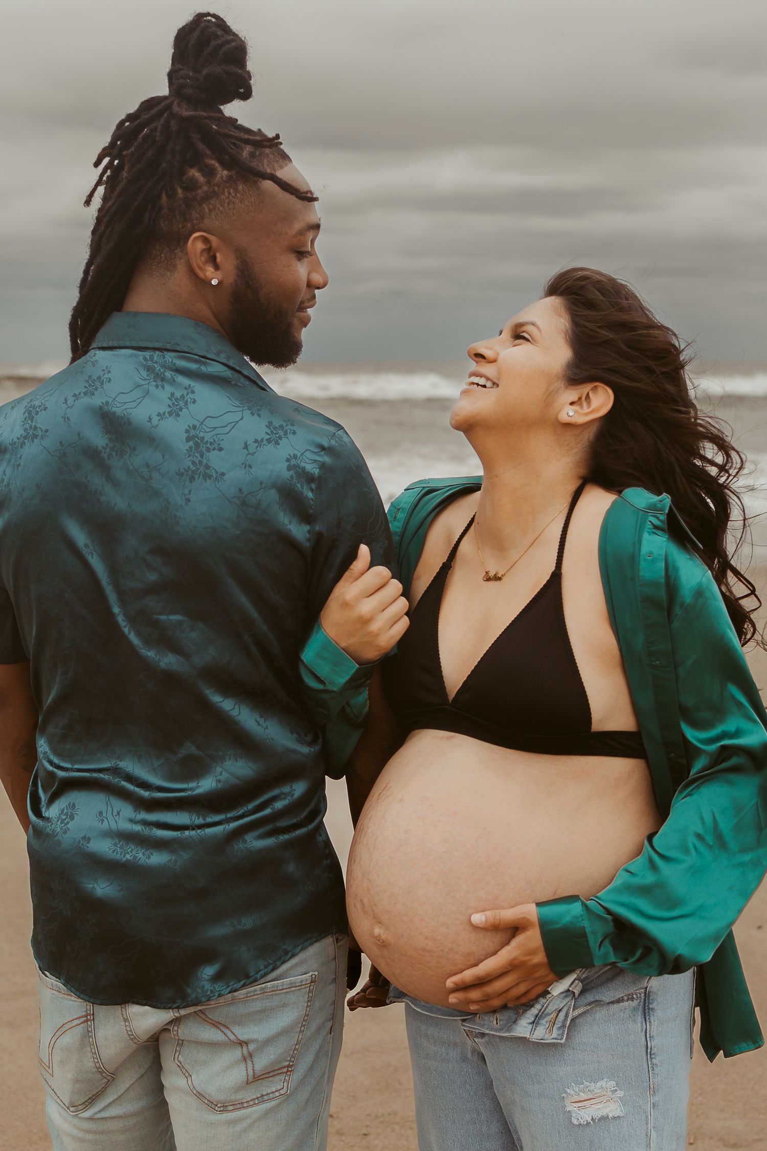 Interracial couple on stormy beach, pregnant woman in black bikini top and green jacket smiling at partner.