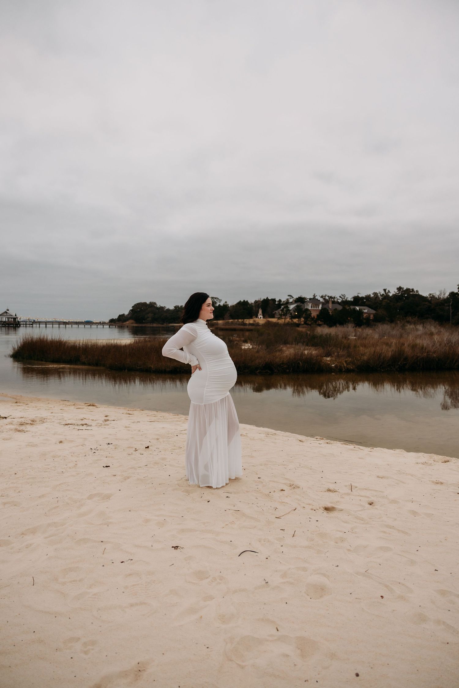 Pregnant woman in white lace gown standing on sandy beach near calm water on overcast day.