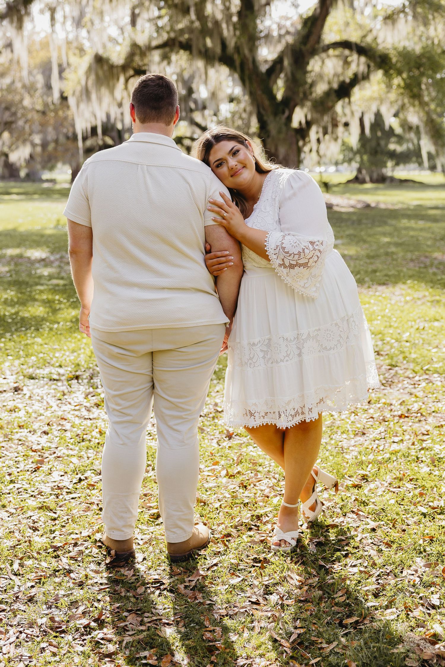 Woman in white lace dress leans on partner's arm while walking through moss-covered park.