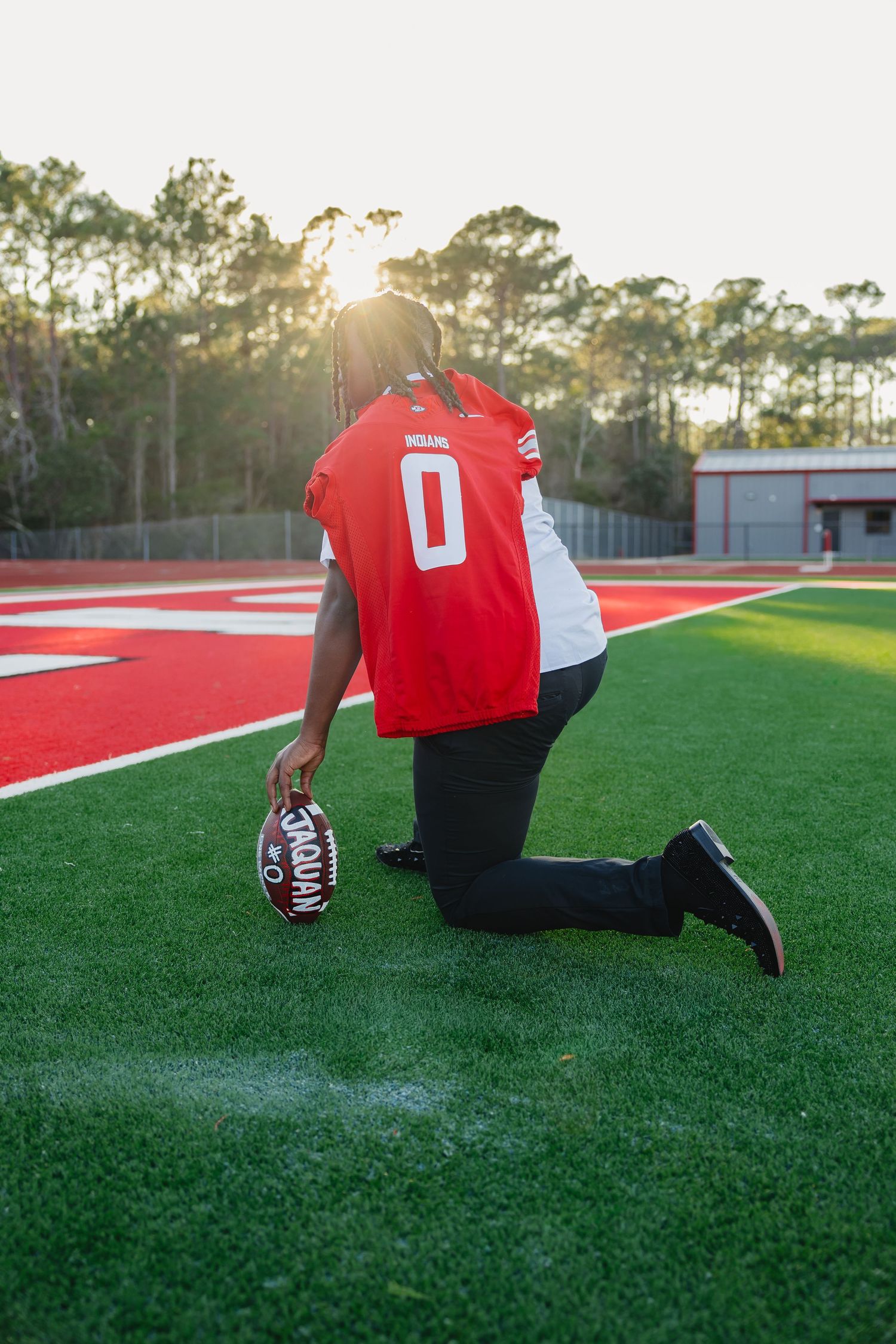 Football player in #0 jersey kneeling on red turf field at sunset holding personalized football.