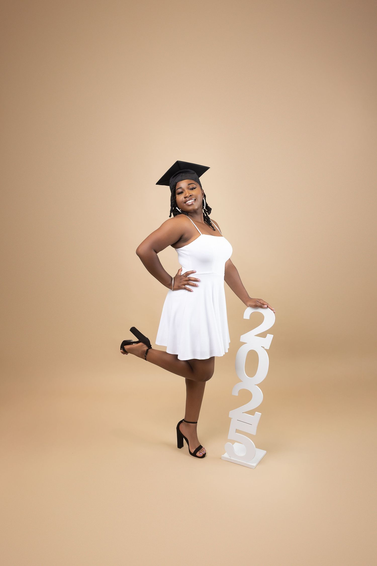 Smiling graduate in white dress and cap posing playfully with 2025 sign on beige background.