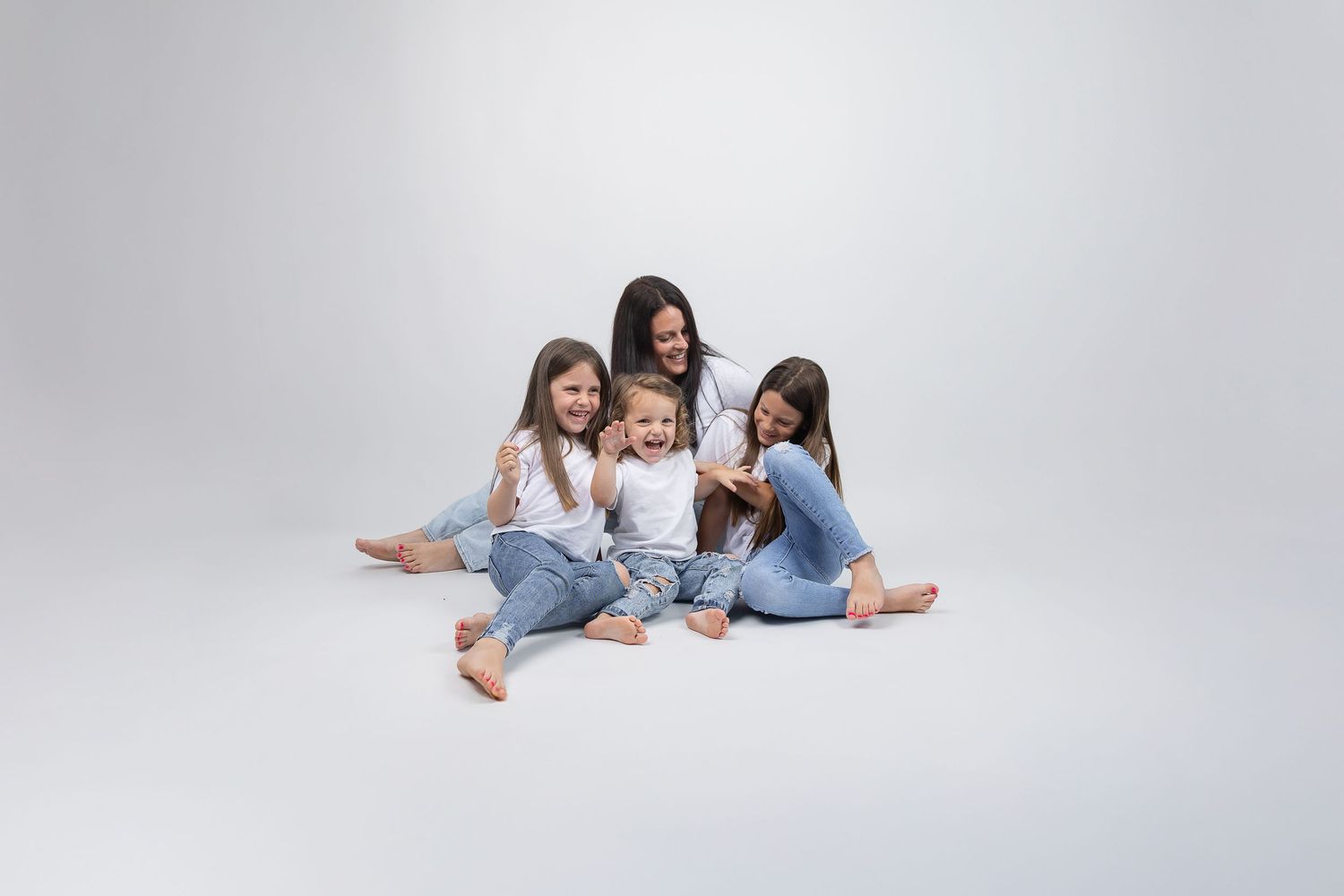 Joyful mother and three daughters in white tops and jeans laugh together on white studio background.