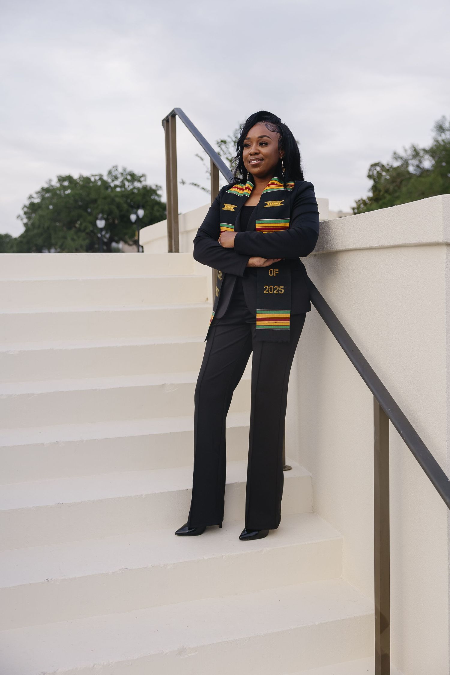 A smiling graduate in black outfit with kente stole reading 2025 poses on white outdoor stairs.