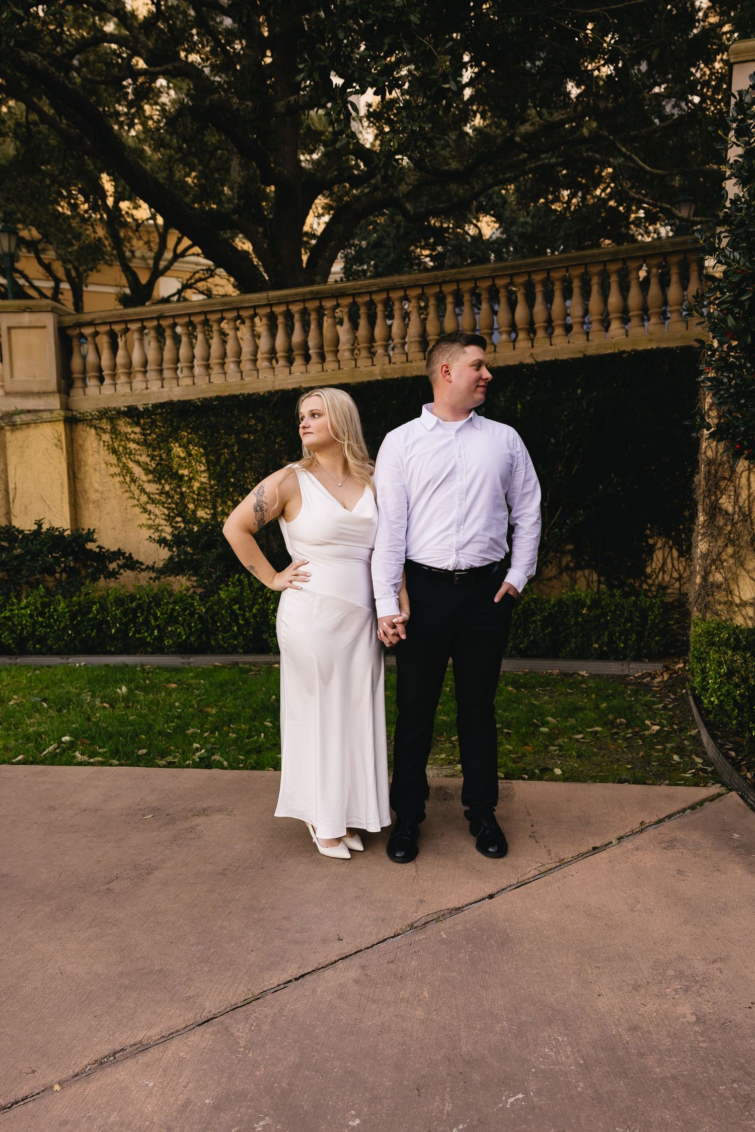 Engaged couple holding hands in white dress and black pants standing before stone balustrade at golden hour.