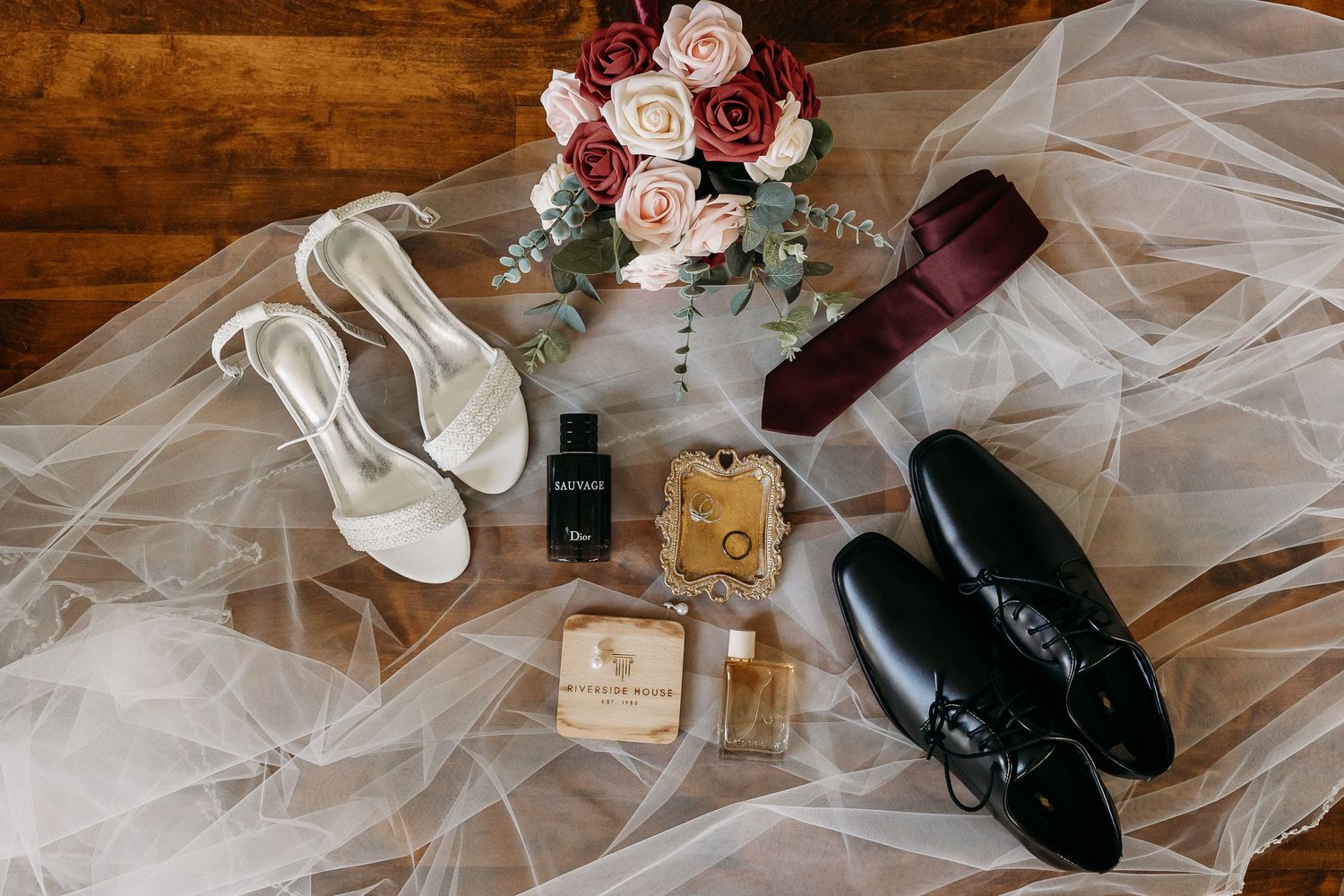 Flat lay of wedding details including white heels, black dress shoes, burgundy tie, roses bouquet, rings, and perfume on veil.