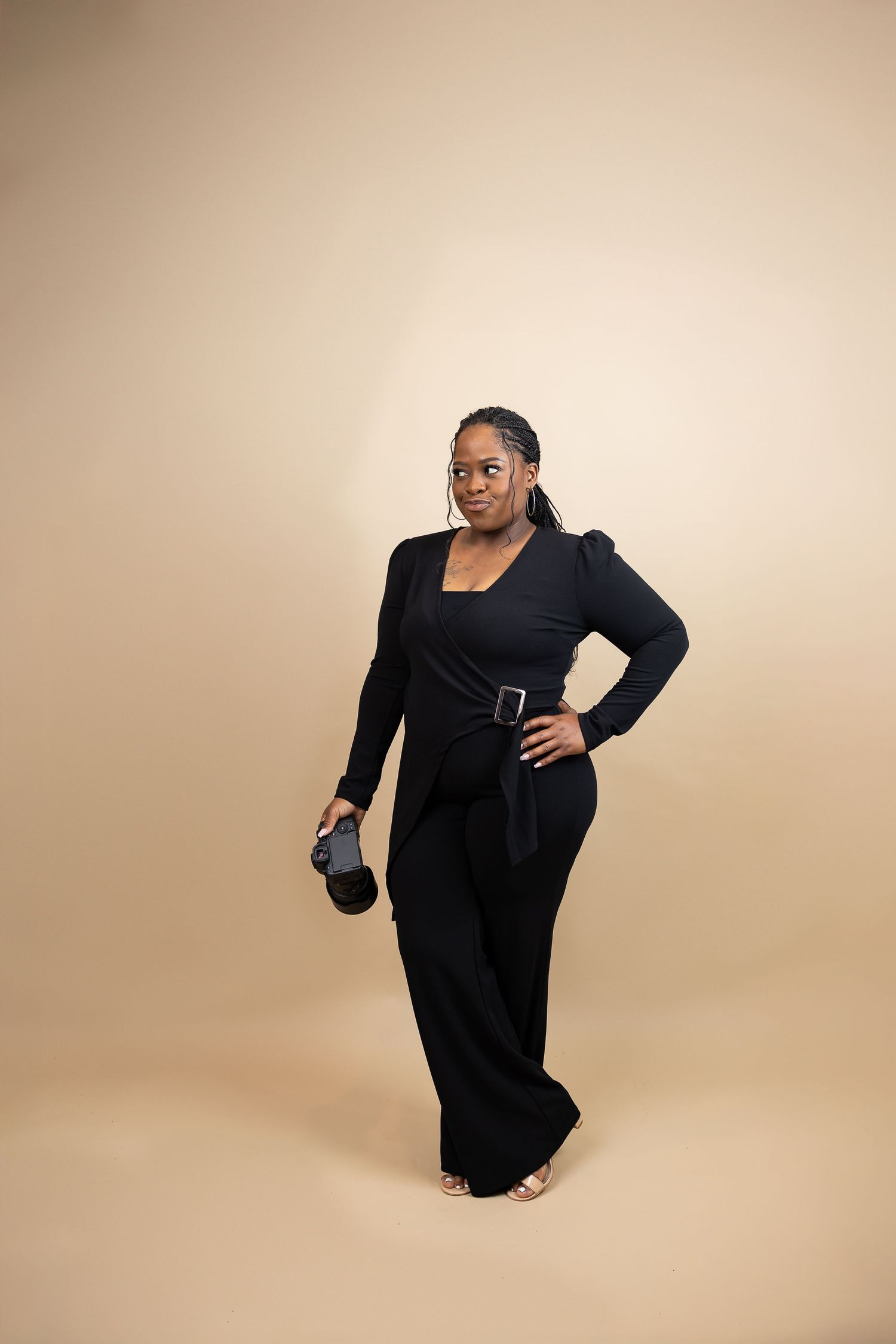 Confident female photographer in black belted jumpsuit holding camera against a beige background.