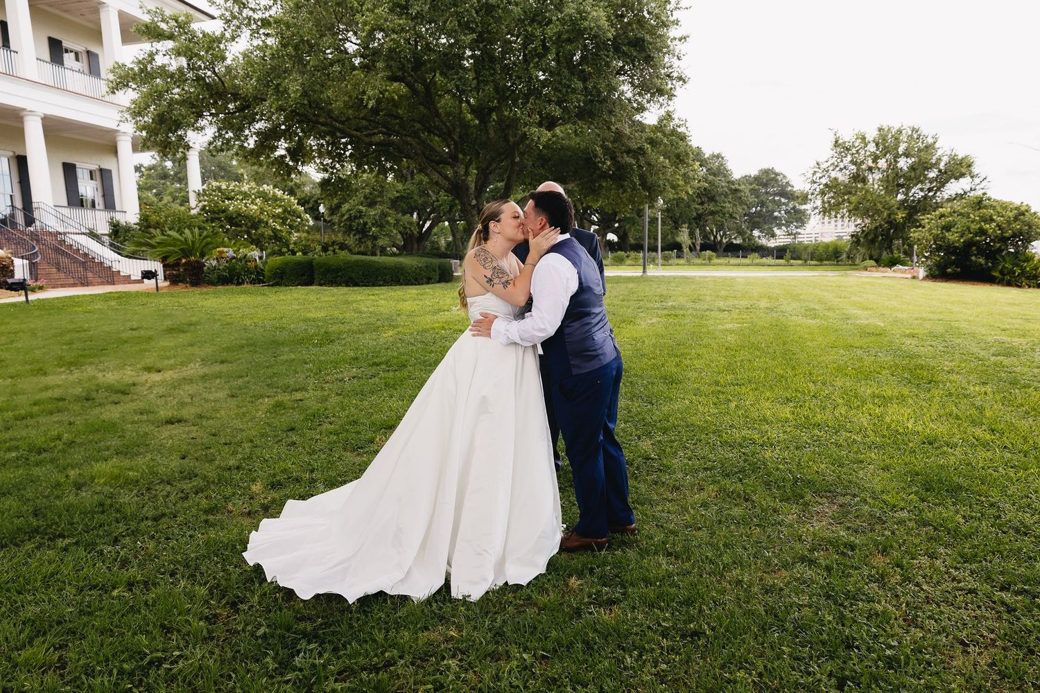 Bride and groom sharing a kiss on lush green lawn in front of elegant white mansion.