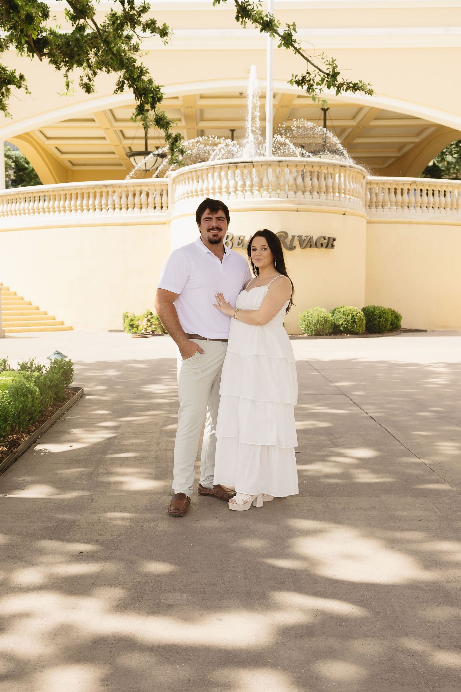 Expecting couple posing in white outfits in front of the Beau Rivage fountain and entrance.