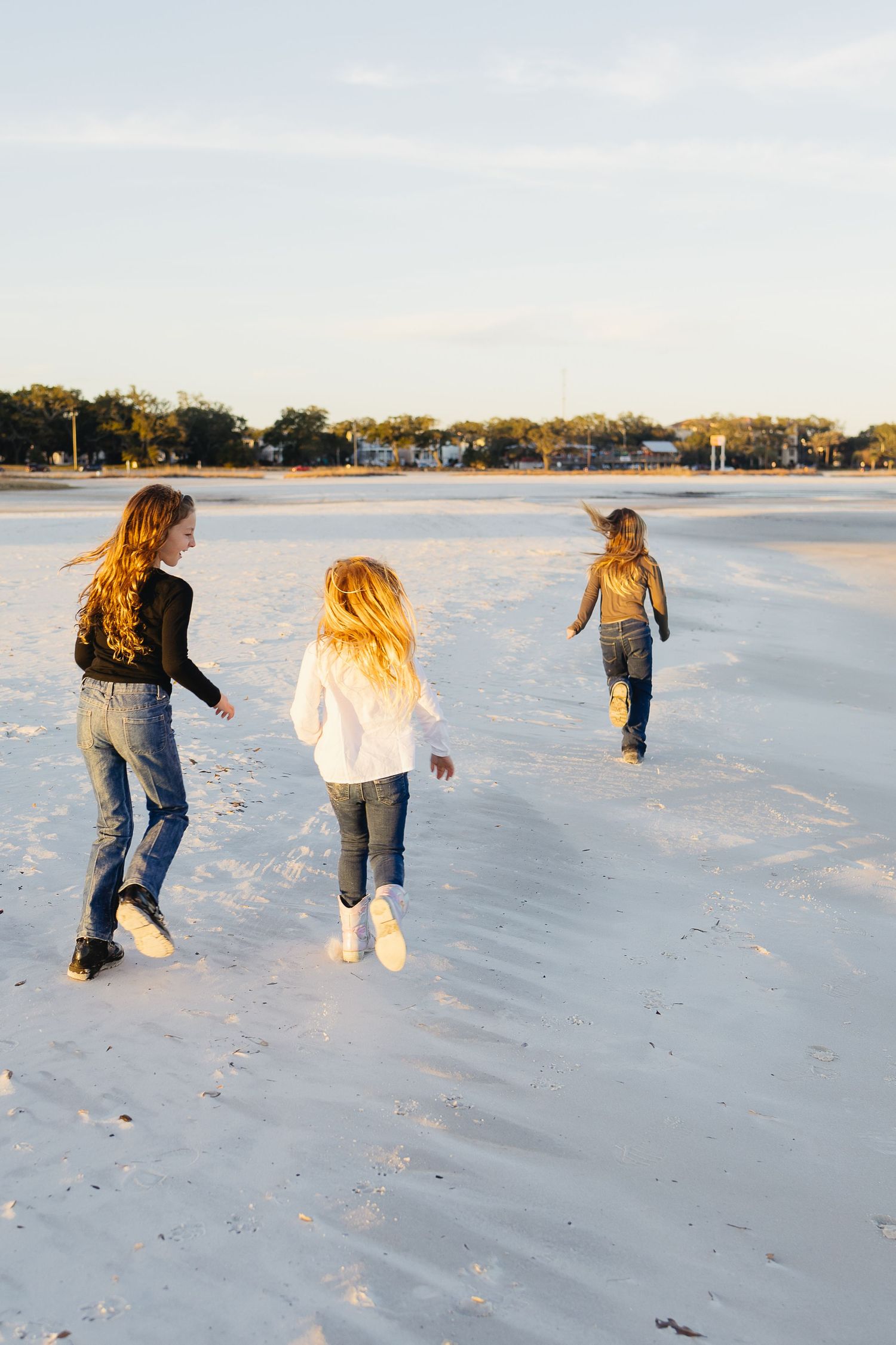Three girls with long hair run and play on a bright sandy beach at golden hour.