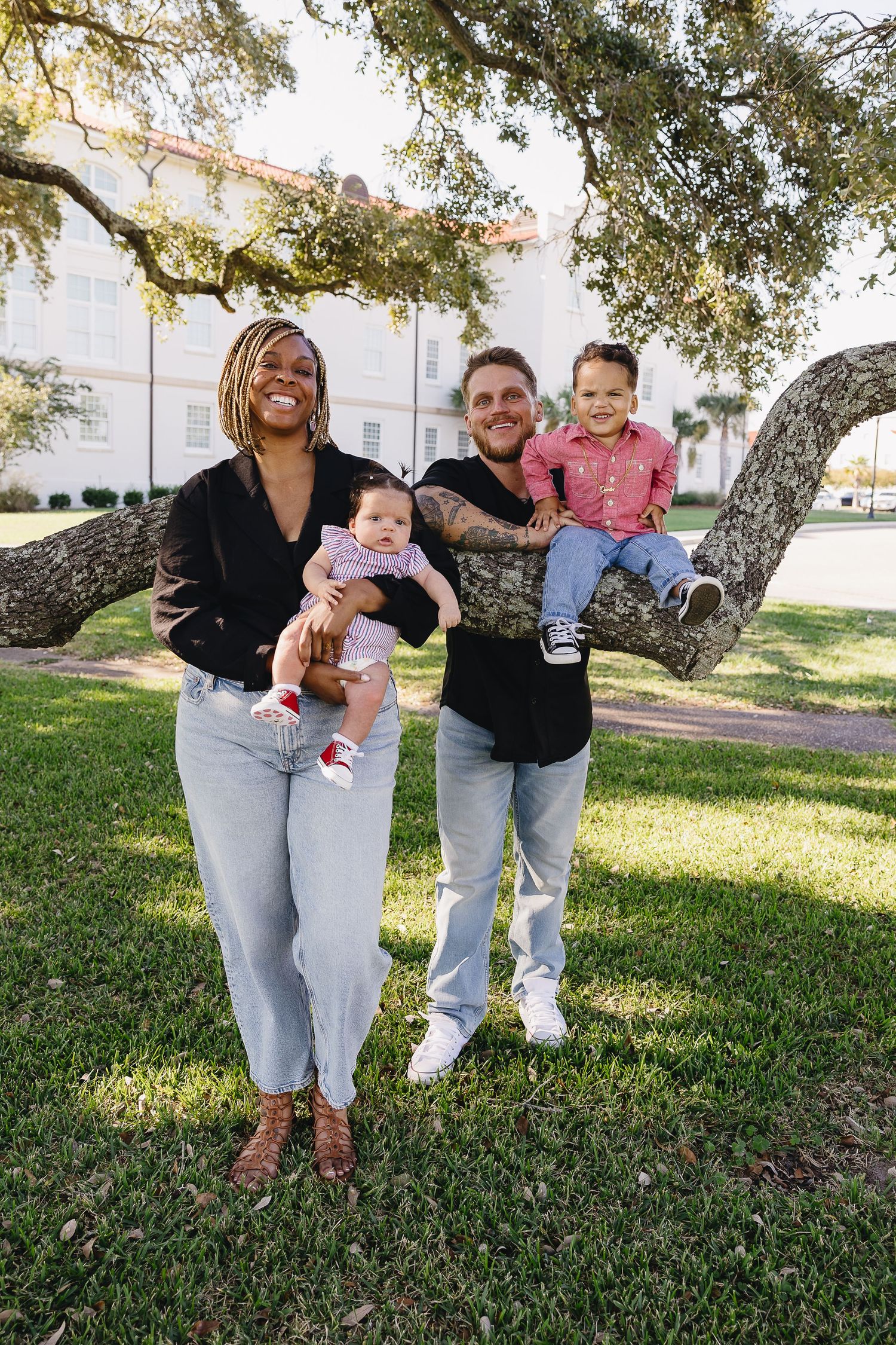 Happy interracial family of four with two young children posing under oak tree on green lawn.