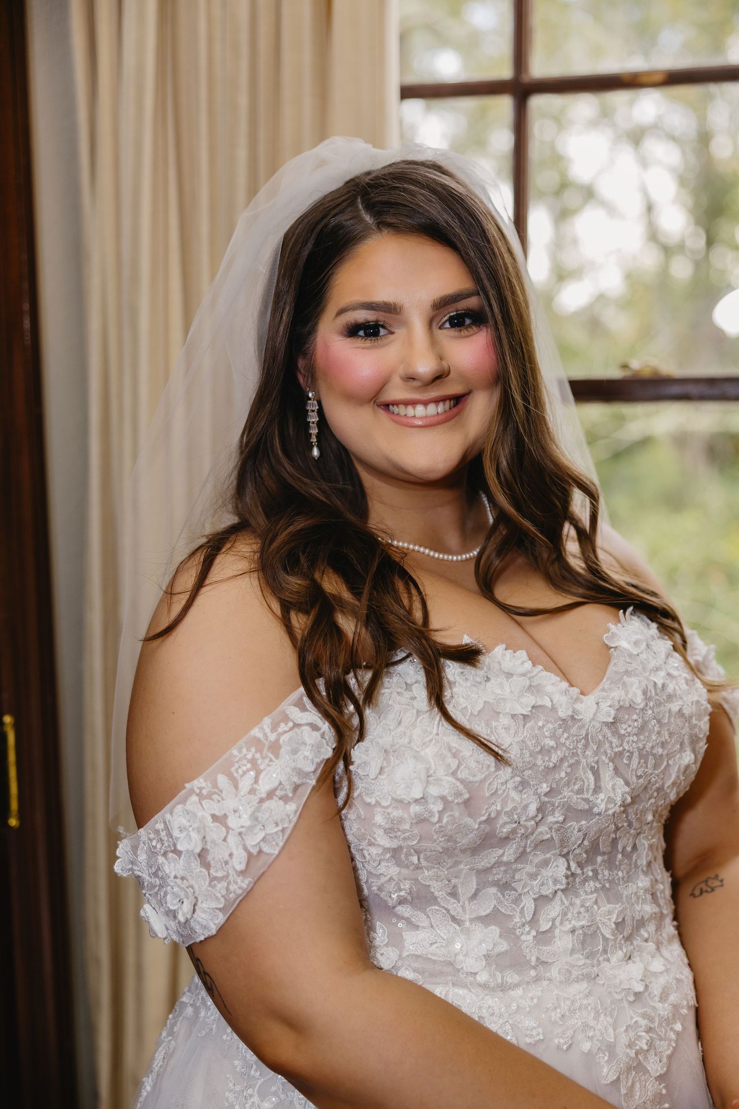 Smiling bride in off-shoulder lace gown with veil and pearl necklace poses by a window.