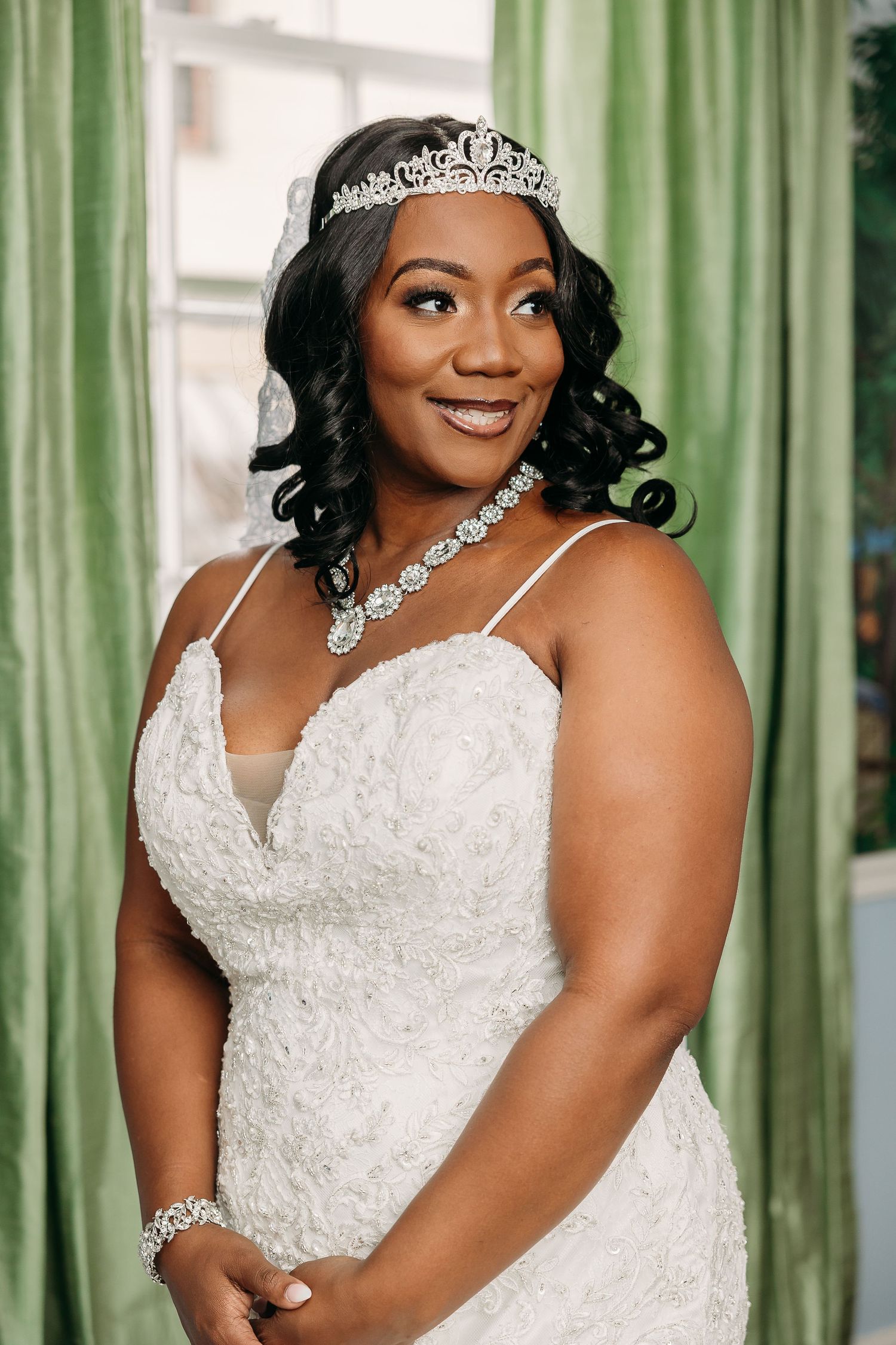Radiant Black bride smiling in beaded lace gown, tiara, and diamond jewelry near green drapes.