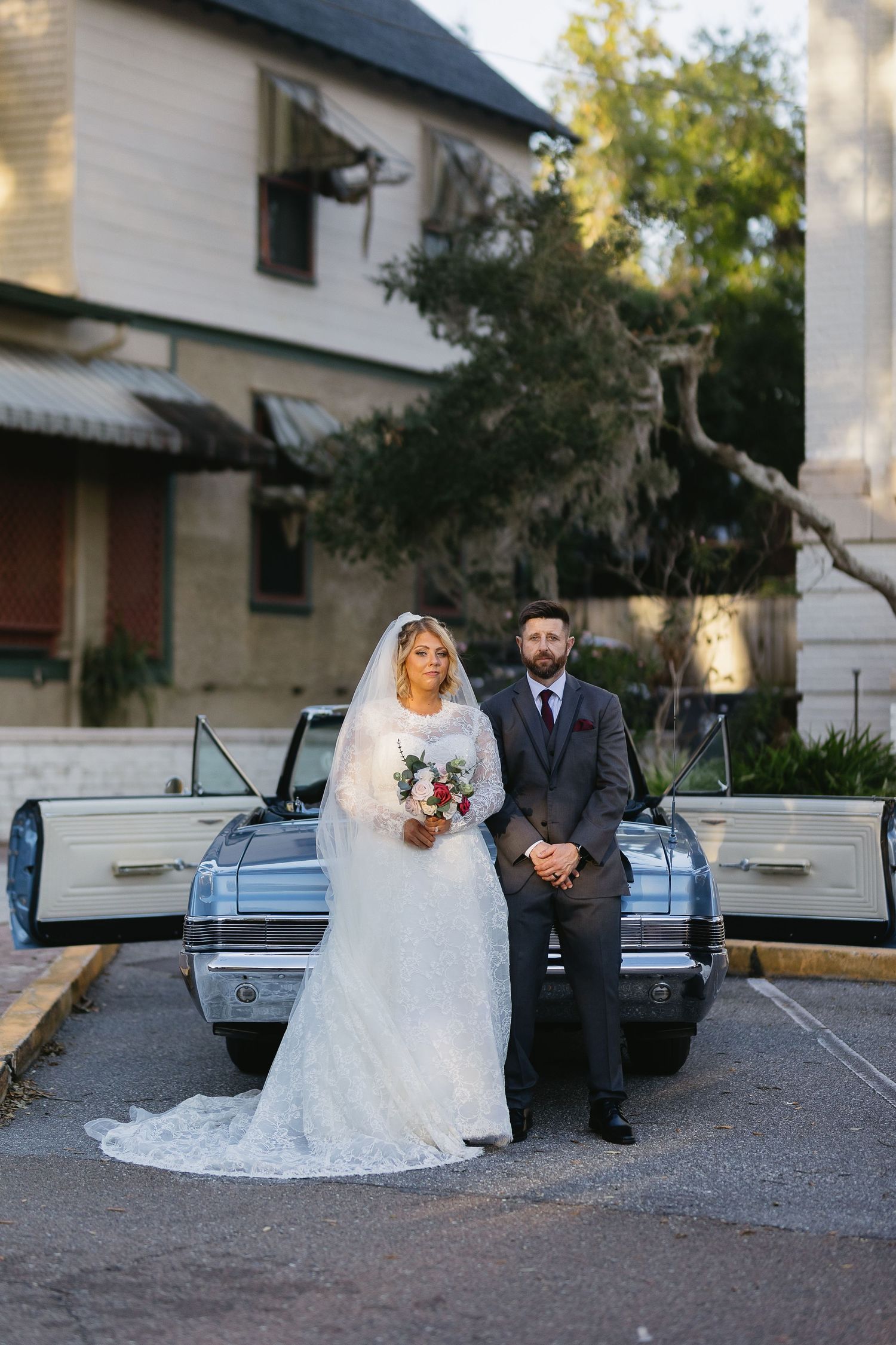 Bride and groom posing together in front of vintage blue convertible at dusk.