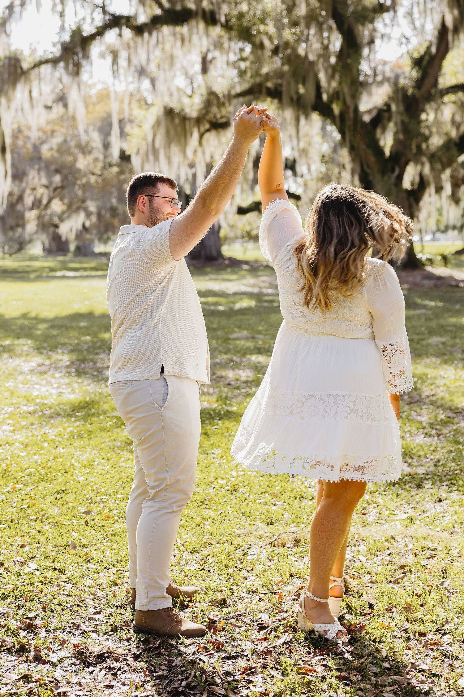 Man twirling woman in white lace dress under beautiful Spanish moss-covered oak trees.