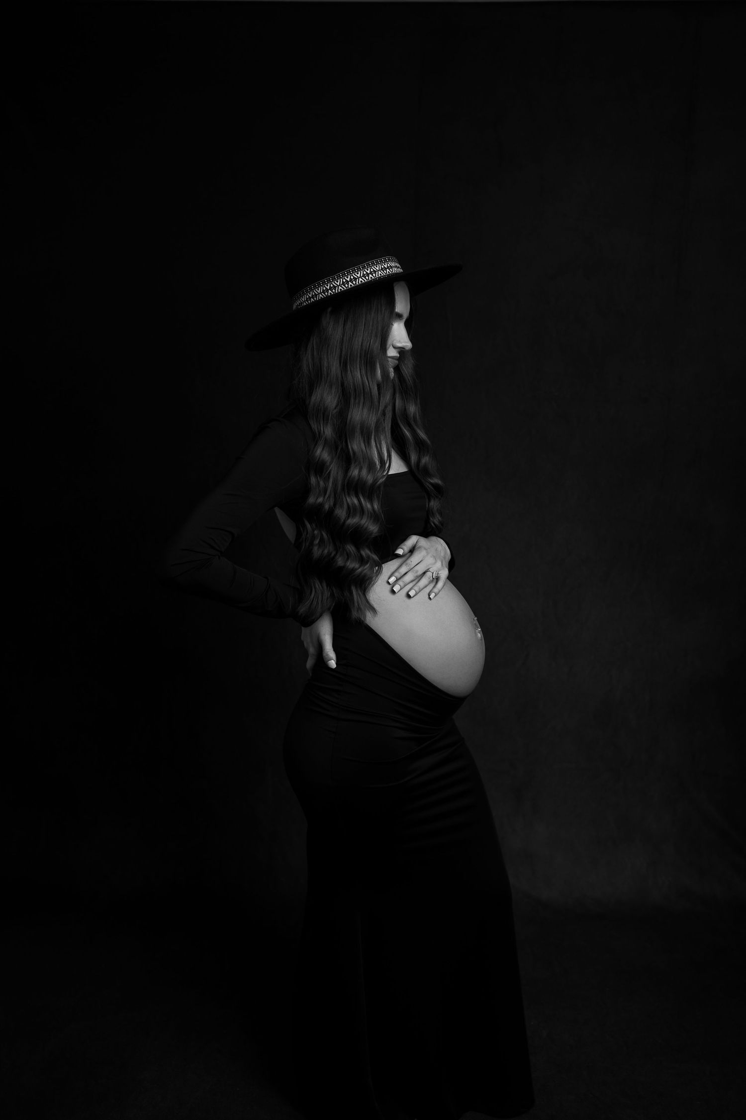 Black and white maternity portrait of pregnant woman in black dress wearing a wide-brim hat.