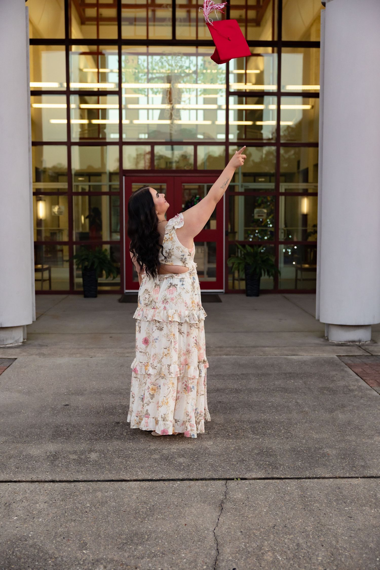 Graduate in floral dress tossing red 2026 cap outside school building entrance.