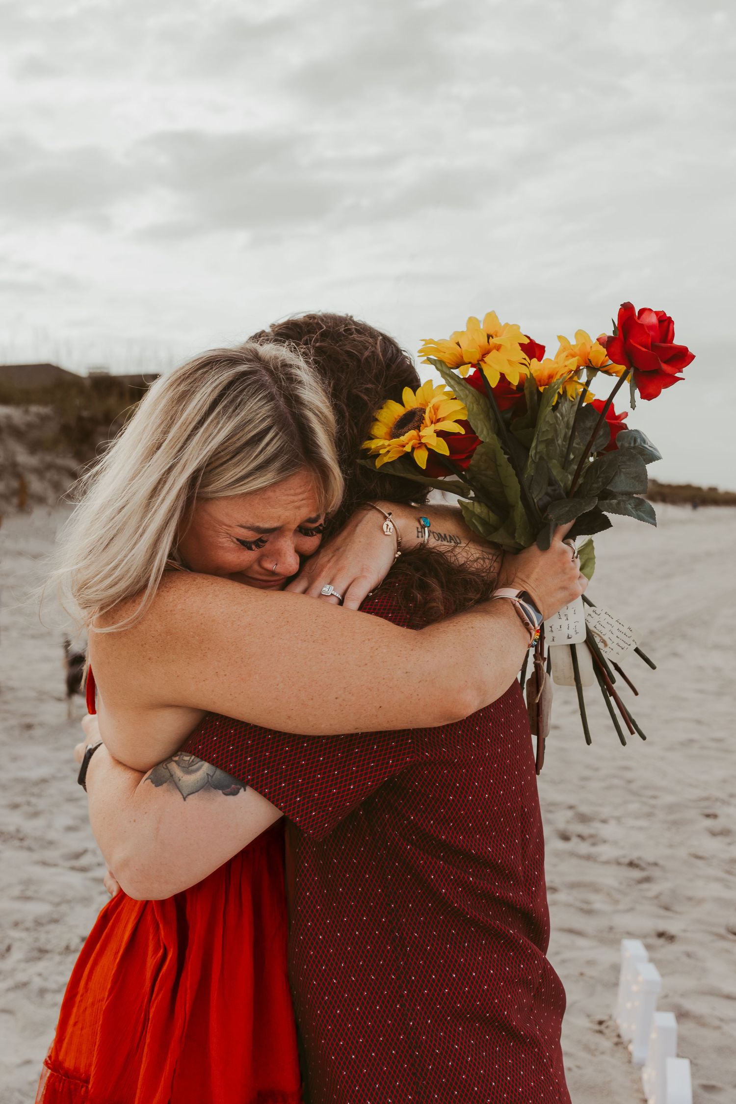 Two women embrace emotionally on beach after proposal, one holding colorful sunflower and rose bouquet.