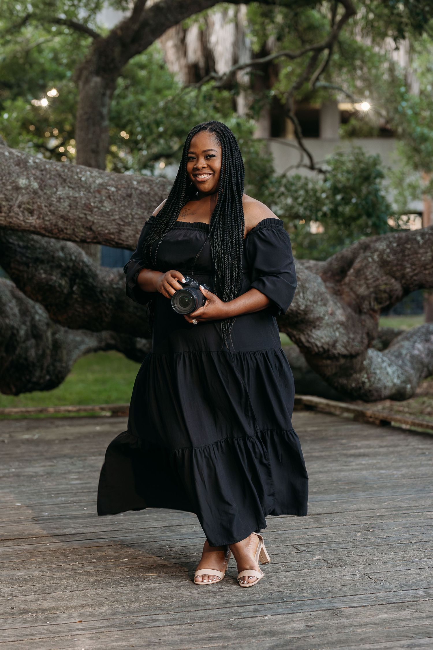 A smiling woman in a black off-shoulder tiered maxi dress holds a camera outdoors near a large tree.