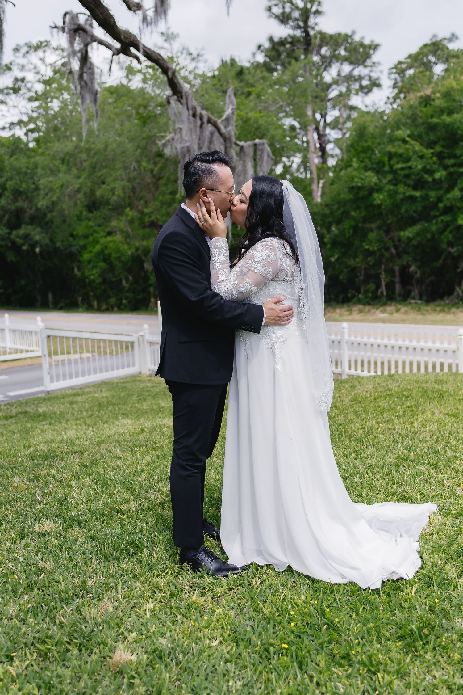 Bride and groom sharing first kiss on green lawn with white fence backdrop.