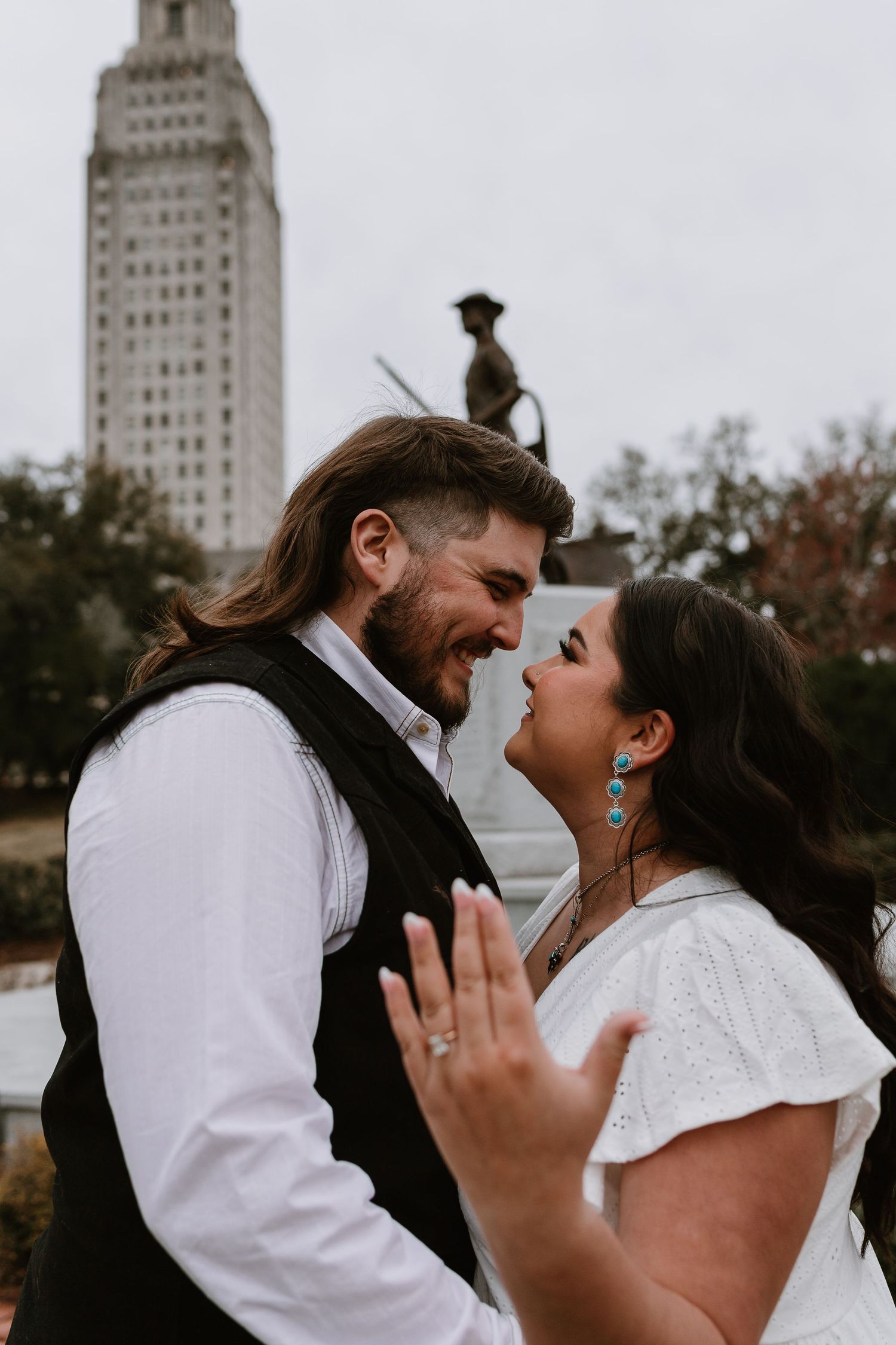 Smiling couple shares joyful moment with groom in black vest, bride in white dress showing engagement ring near capitol.