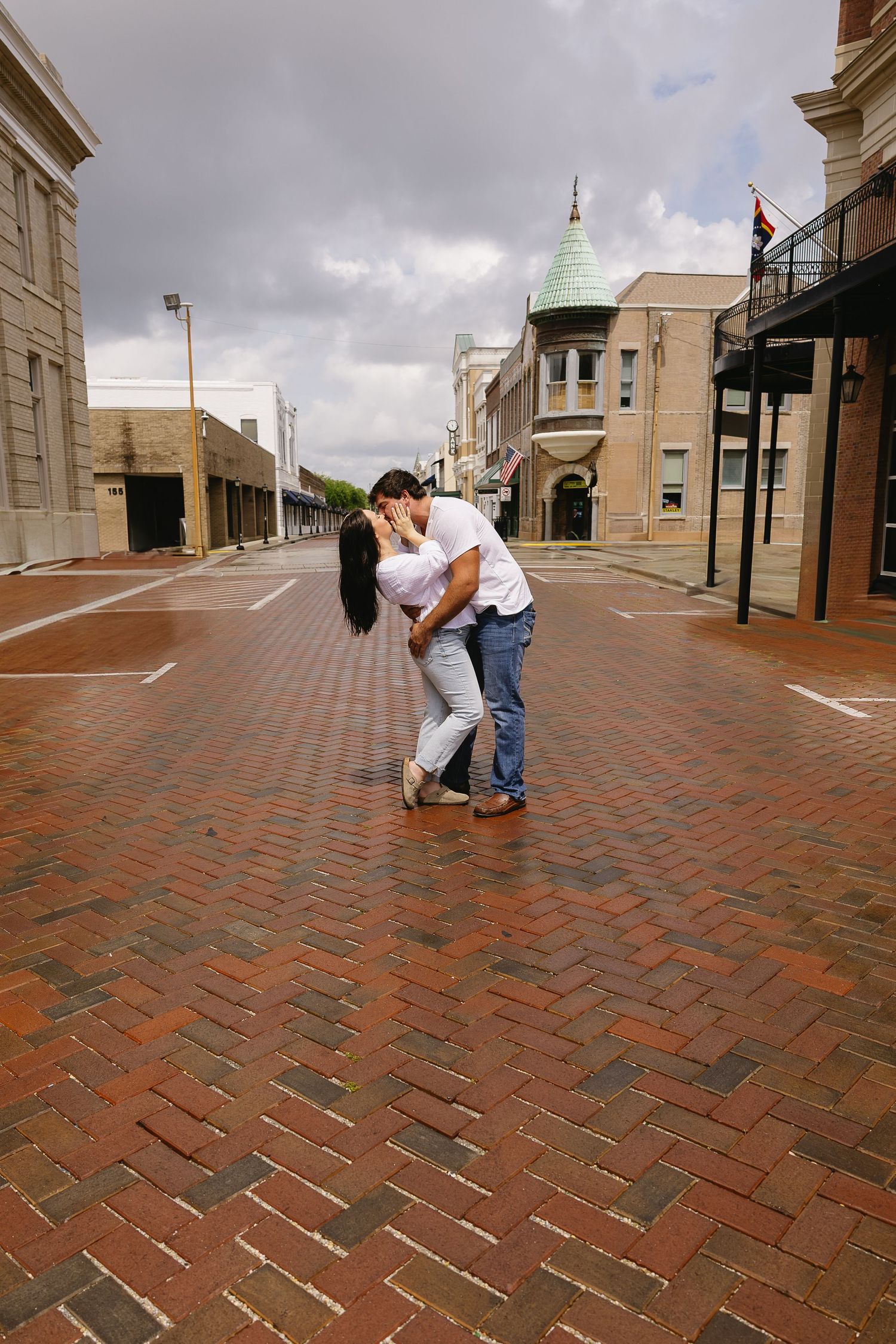 Couple sharing a romantic kiss on a brick street with a green-domed building in the background.