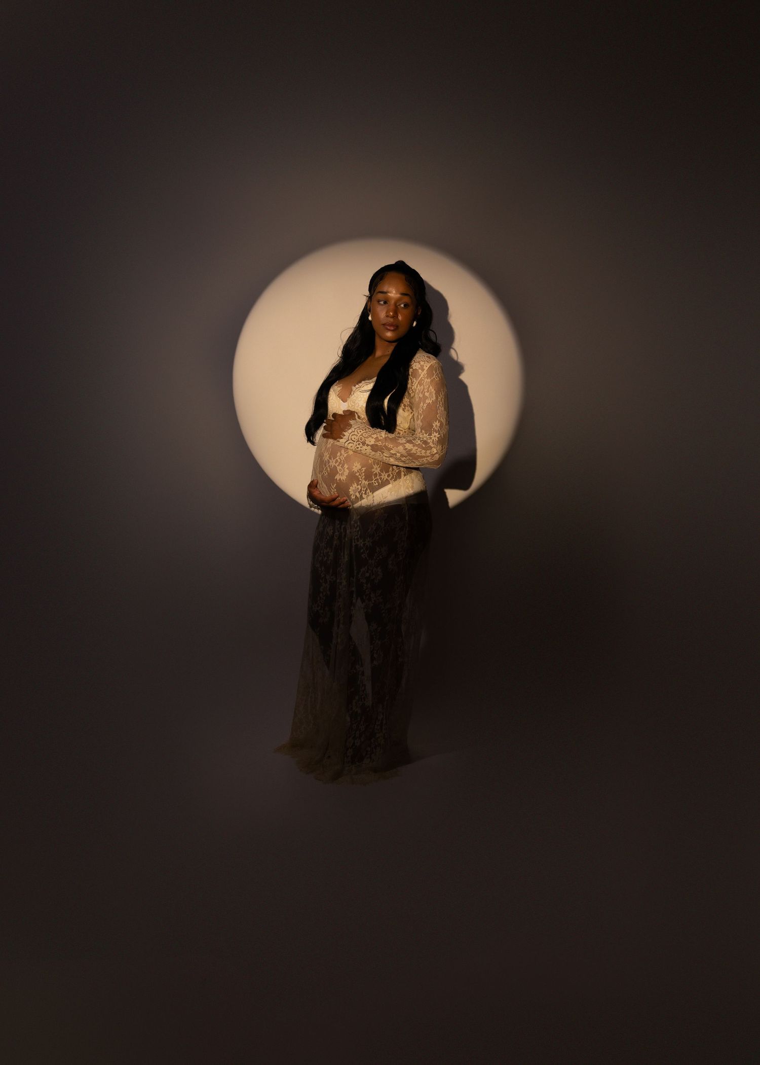 Pregnant Black woman in lace gown stands in dramatic circular spotlight against dark background.