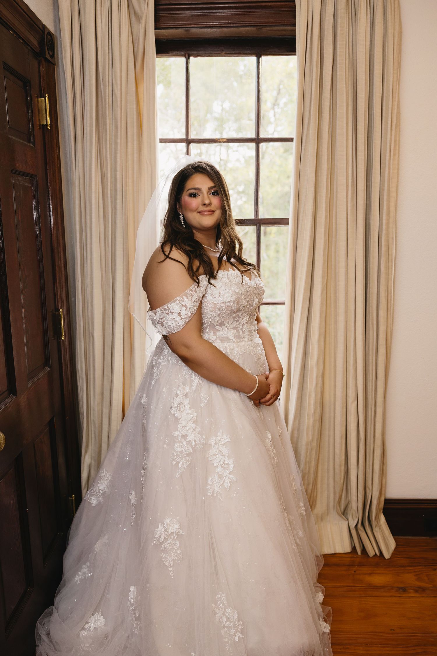 Bride in off-shoulder lace ballgown poses by a large window in an elegant room.