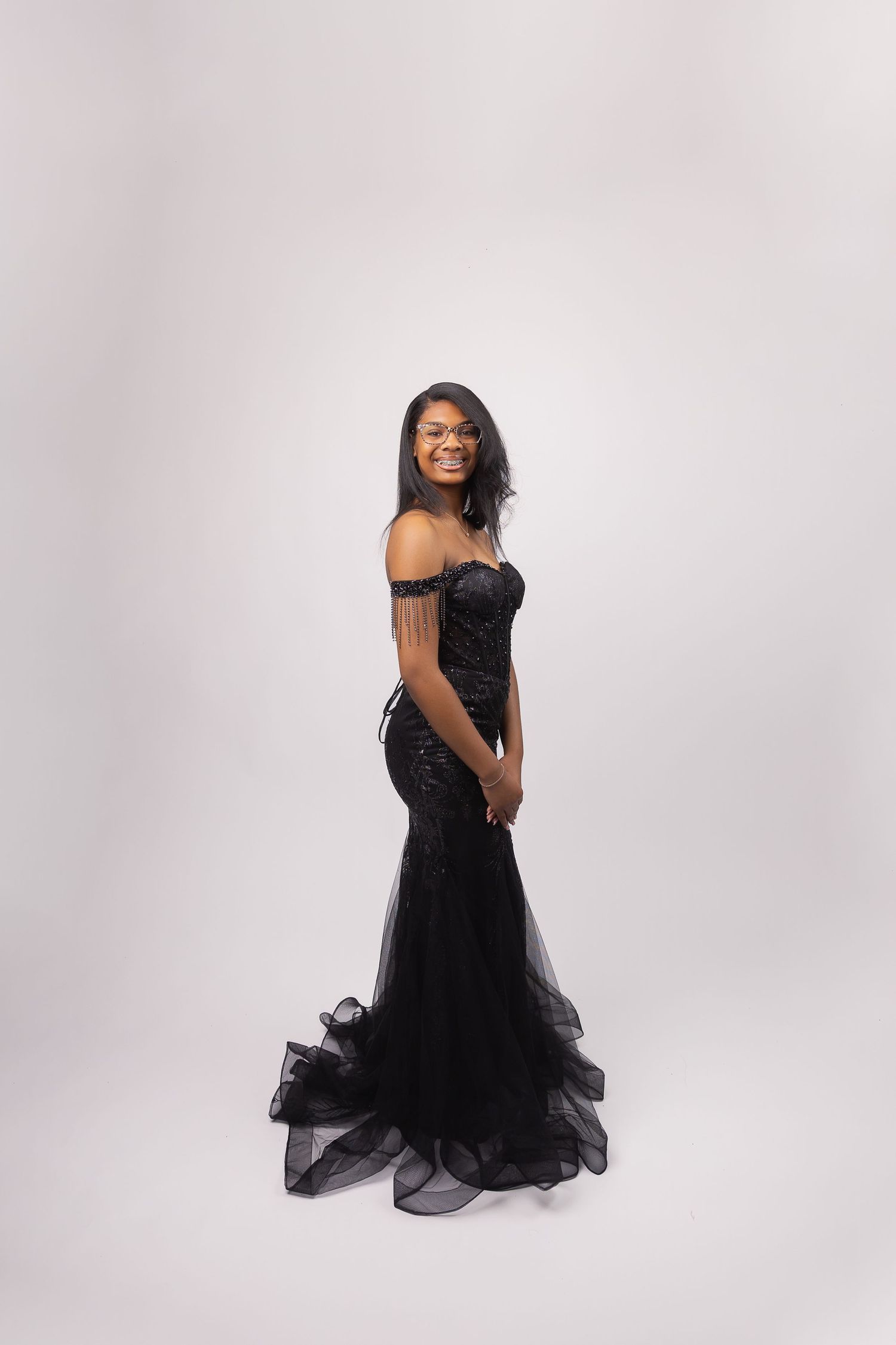 Young woman in elegant black off-shoulder mermaid gown smiling over shoulder in studio portrait.