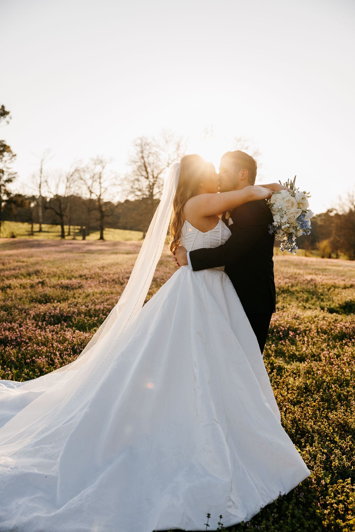 Bride and groom share golden sunset kiss in flower field, veil flowing in breeze.
